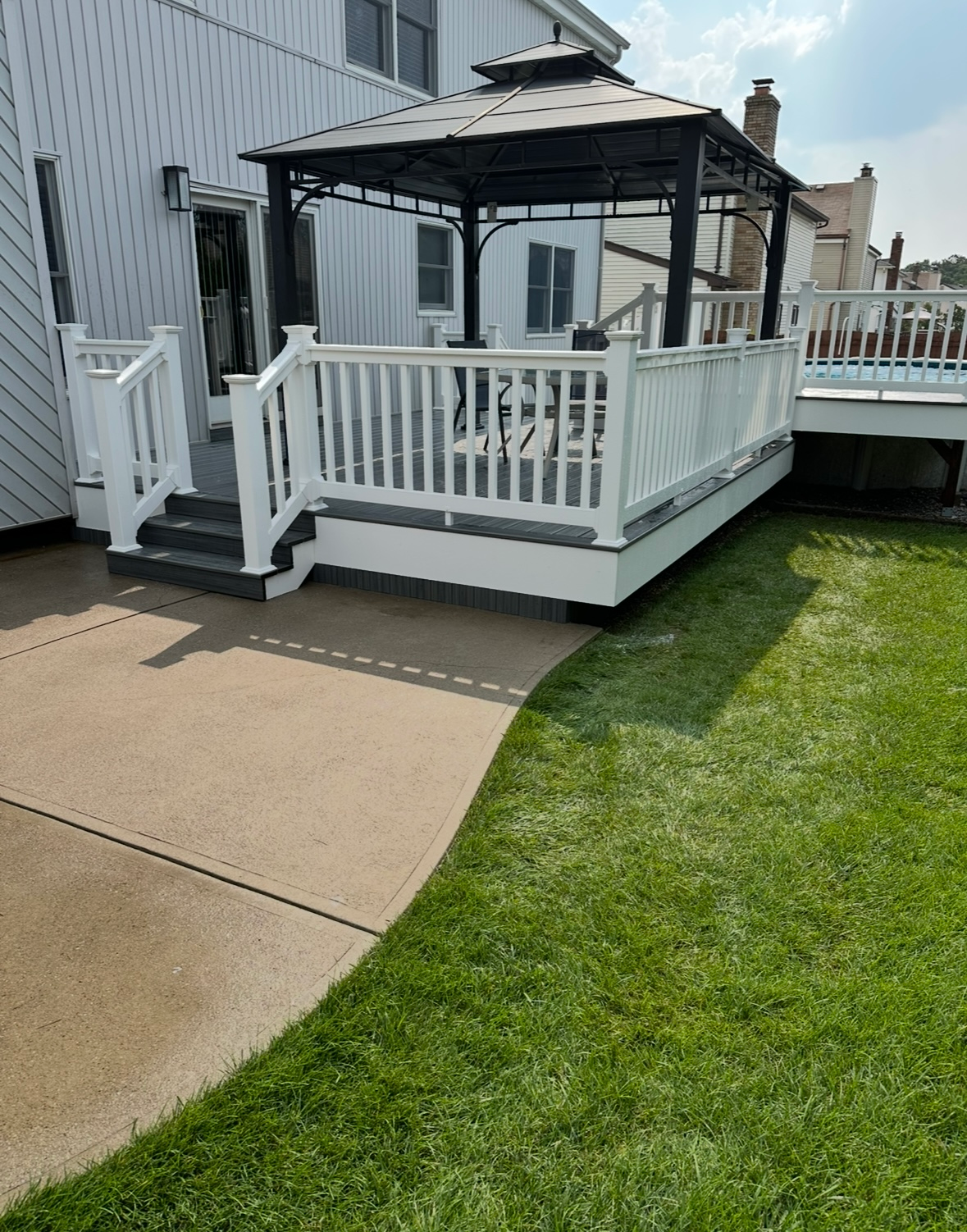 A white deck with a black gazebo, steps, and railings next to a house, situated beside a concrete patio and green lawn.