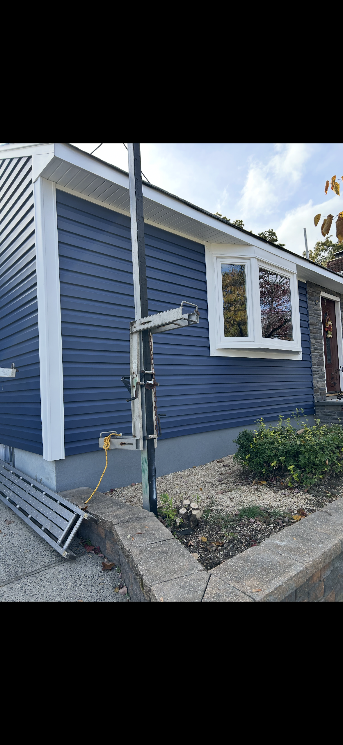 A blue-sided house with white trim and a bay window, featuring an outdoor utility pole and a gravel-filled front yard.