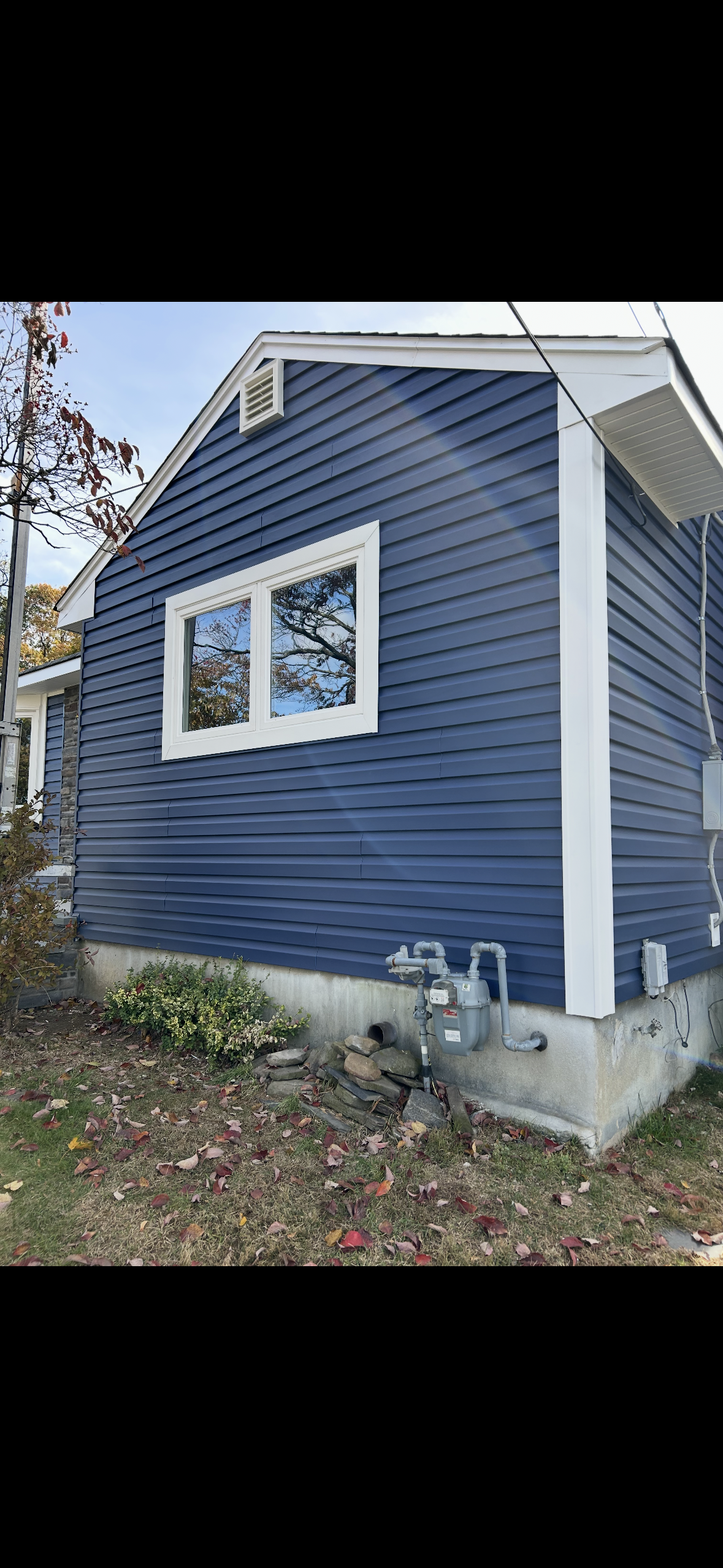 A side view of a house exterior with blue horizontal siding, white trim, a single window, and a gray gas meter near the base.