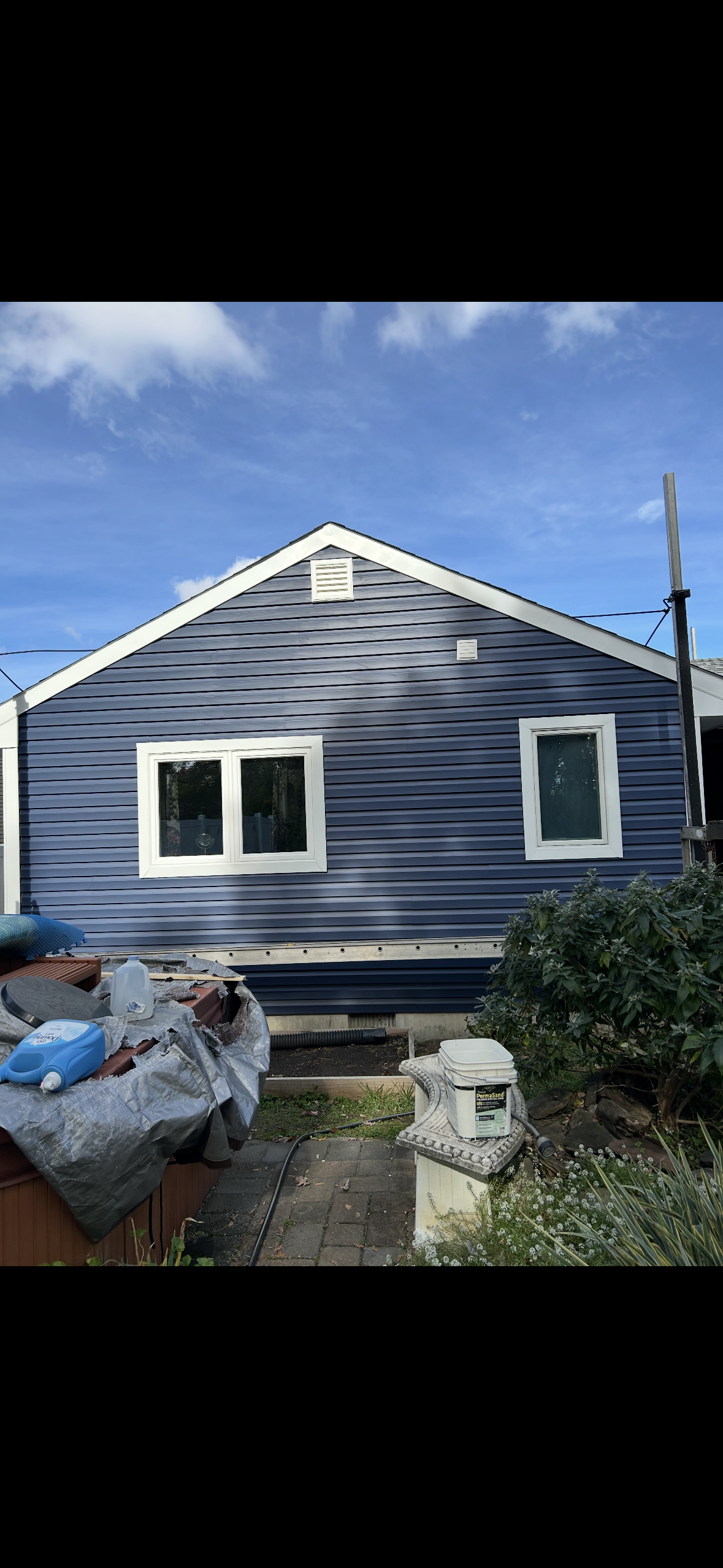 A view of a blue, siding-clad house exterior with two white-framed windows, against a bright, partly cloudy blue sky.