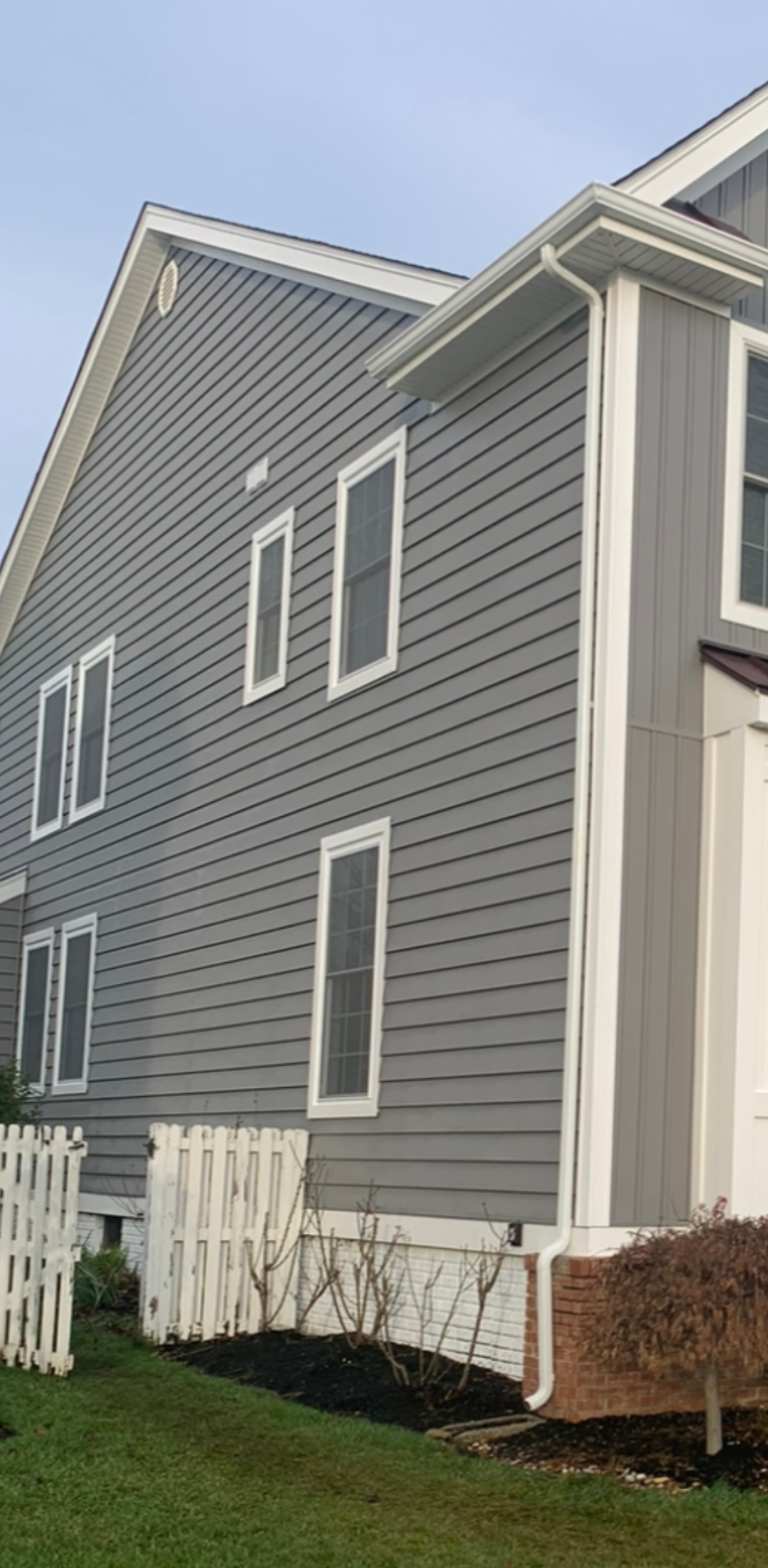 Exterior side view of a gray-sided house with a white picket fence, downspout, and mulched garden bed.