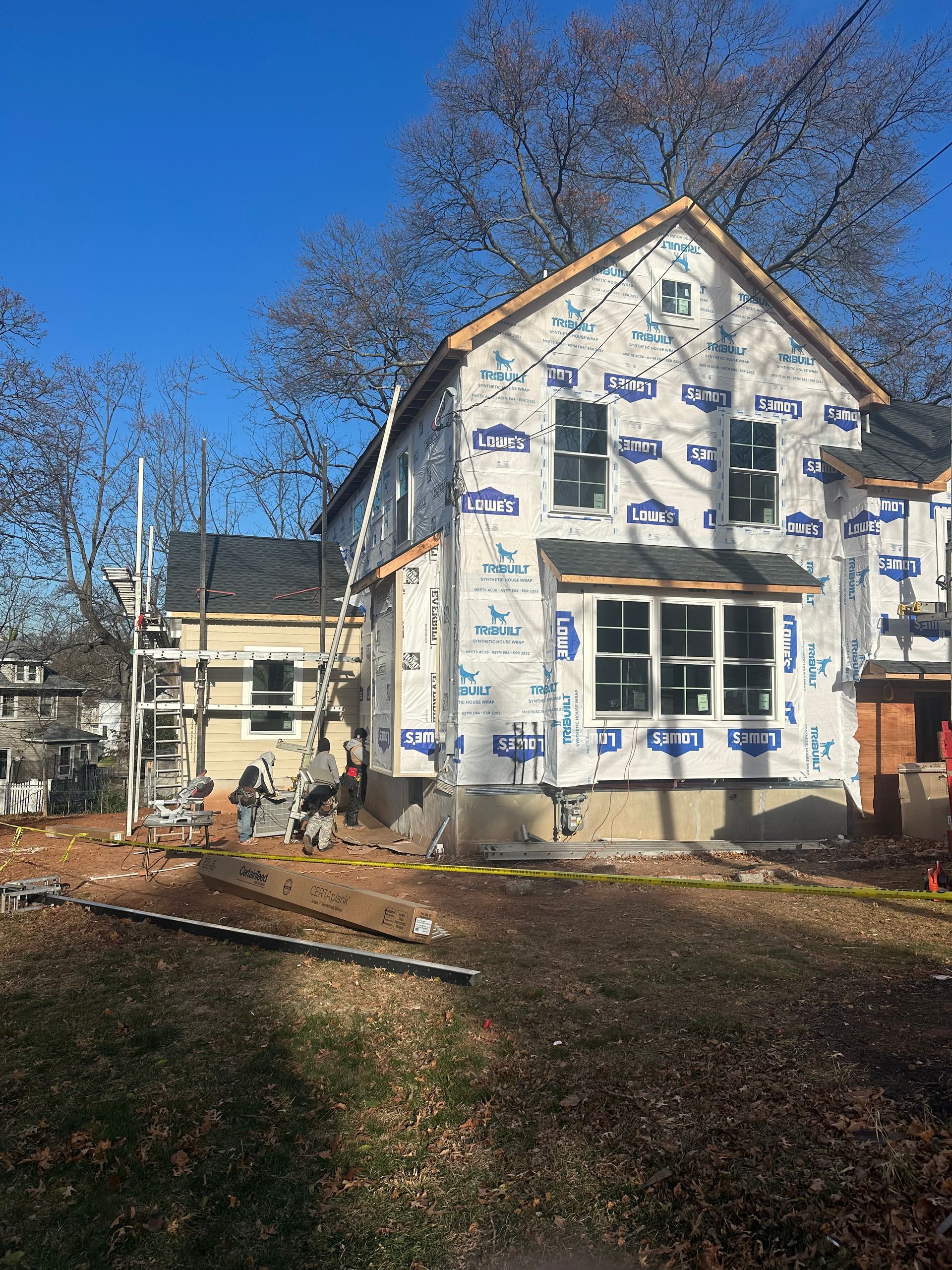 A two-story house under construction, wrapped in white protective sheeting, with scaffolding and a bare yard in autumn.
