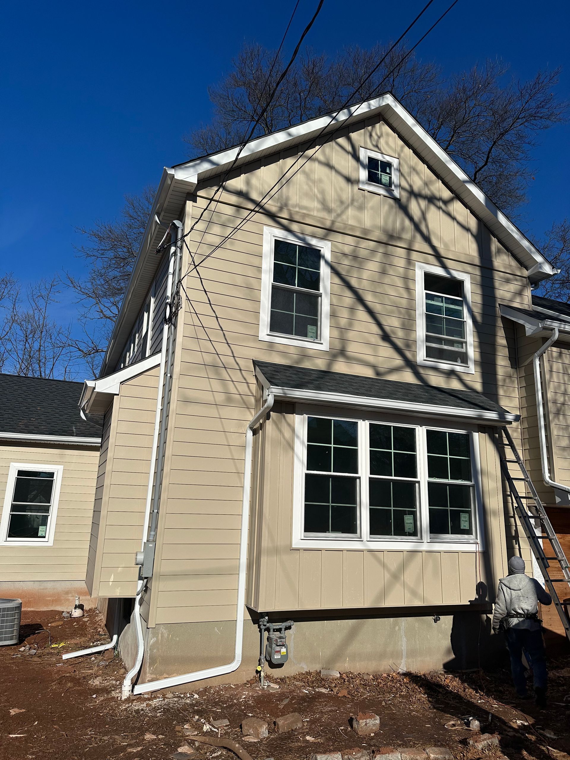 A beige two-story house under construction, featuring new windows, siding being installed, and a ladder on the right.