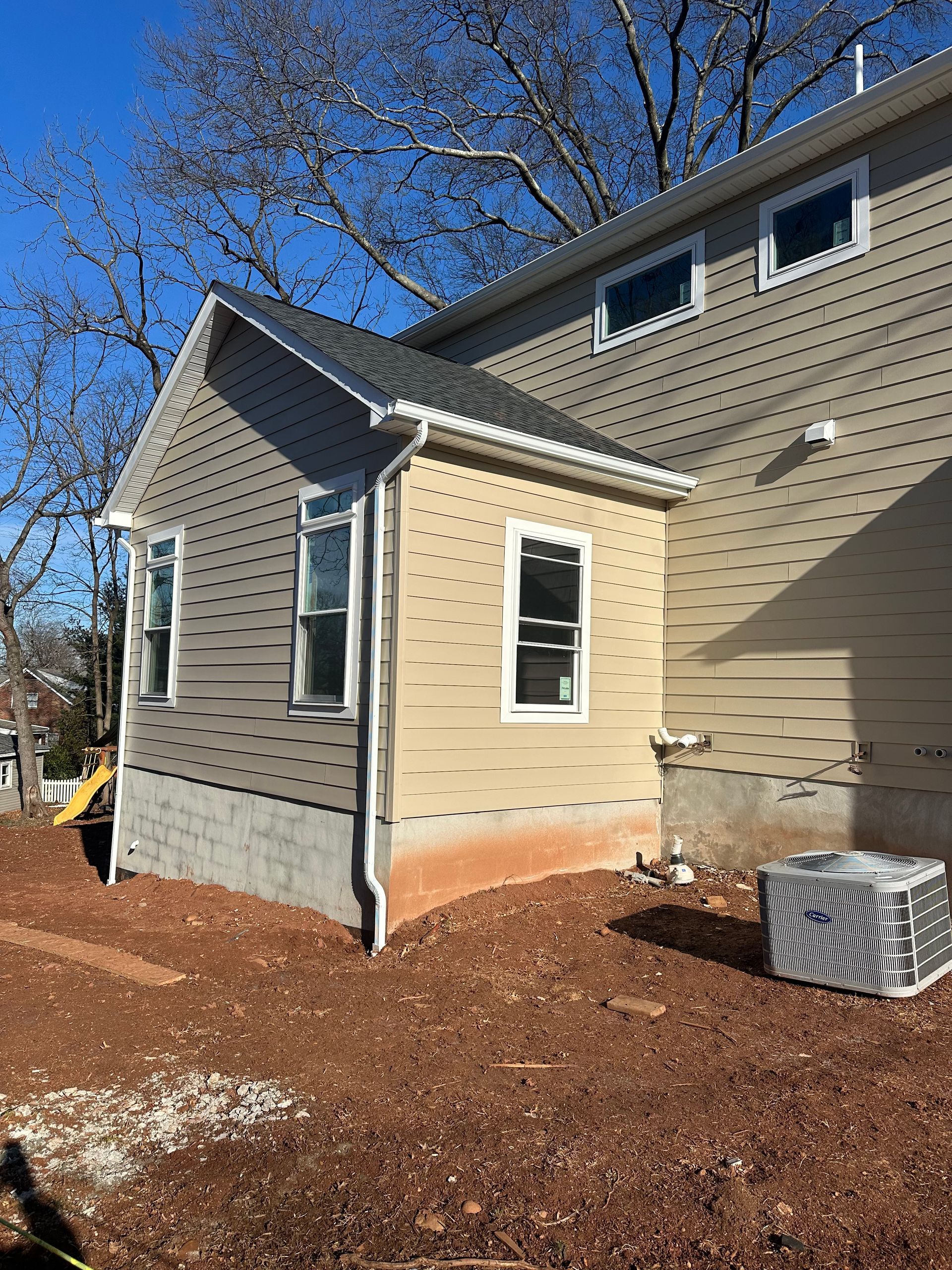 Side view of a tan-sided home addition with new windows, a concrete foundation, and an outdoor air conditioning unit.