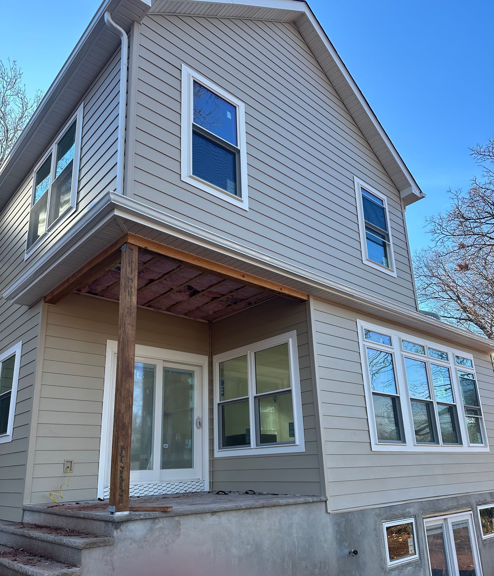 A two-story house exterior under construction with light-colored siding, a covered porch area, and newly installed windows.