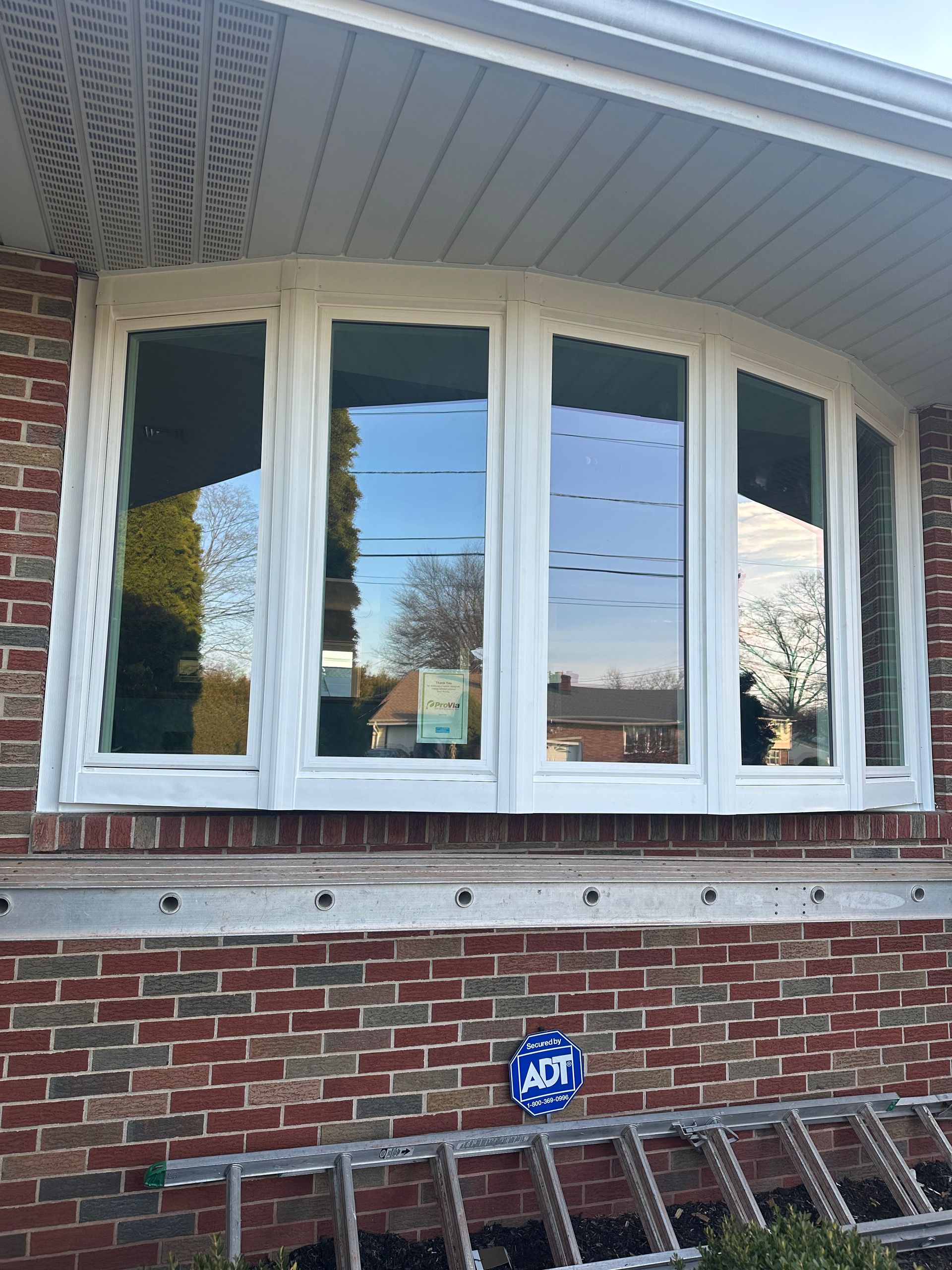 A white bay window installed on a brick exterior wall with a ladder and security sign at the base.
