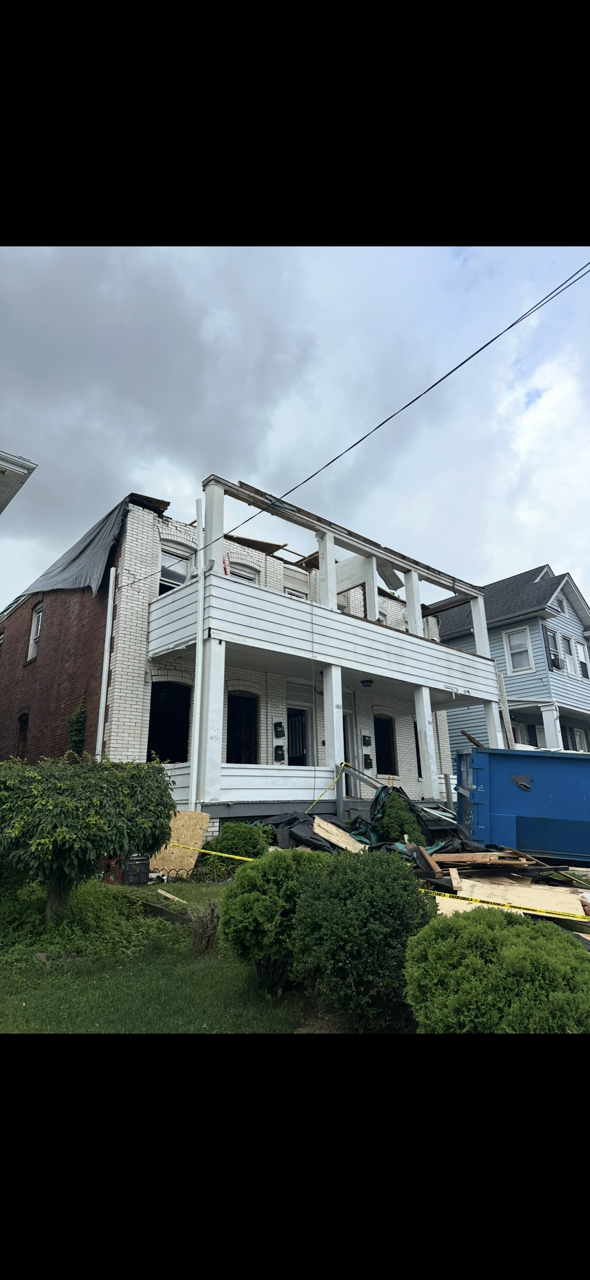 A two-story house under construction with exposed white siding, missing roof sections, and a blue dumpster in the yard.