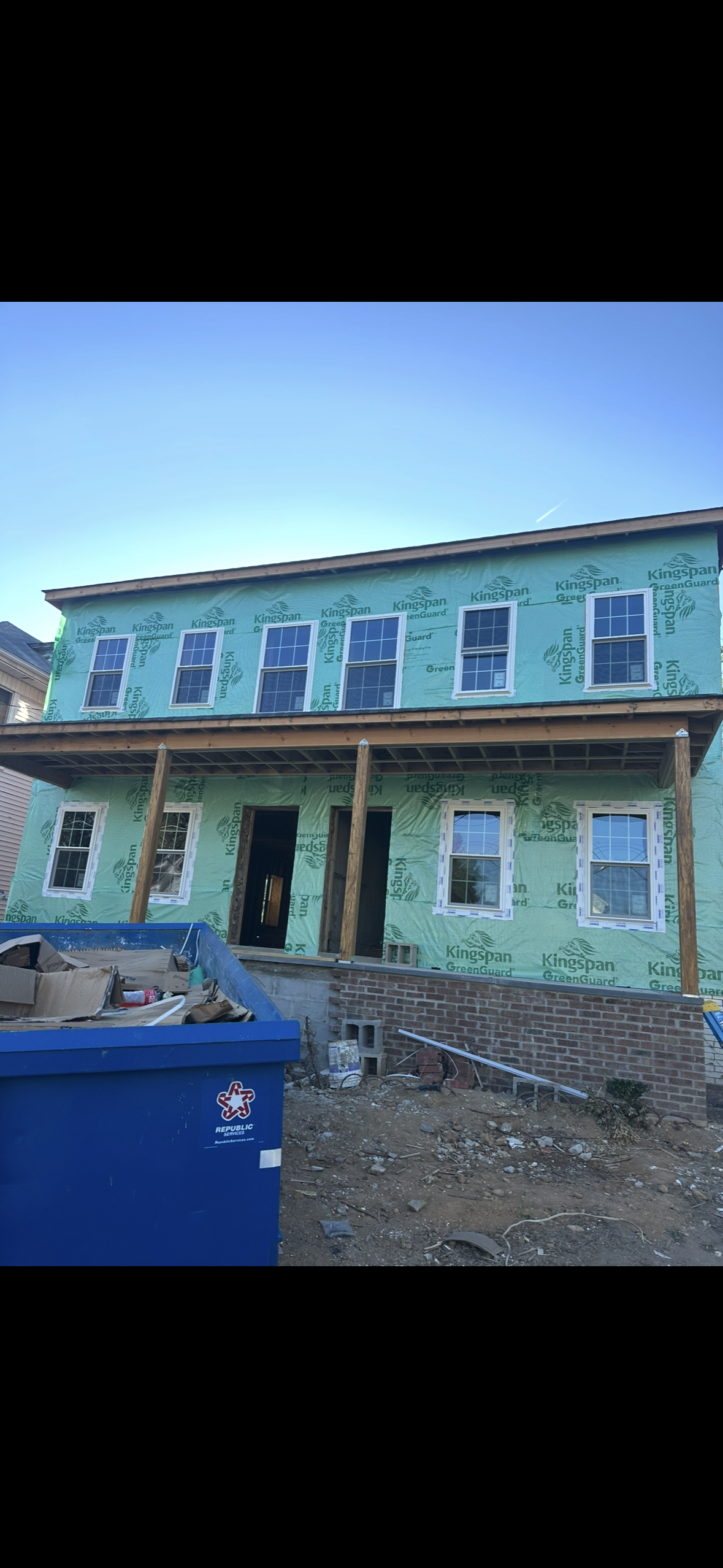 A two-story building under construction with green exterior sheathing, multiple windows, a porch, and a blue dumpster.