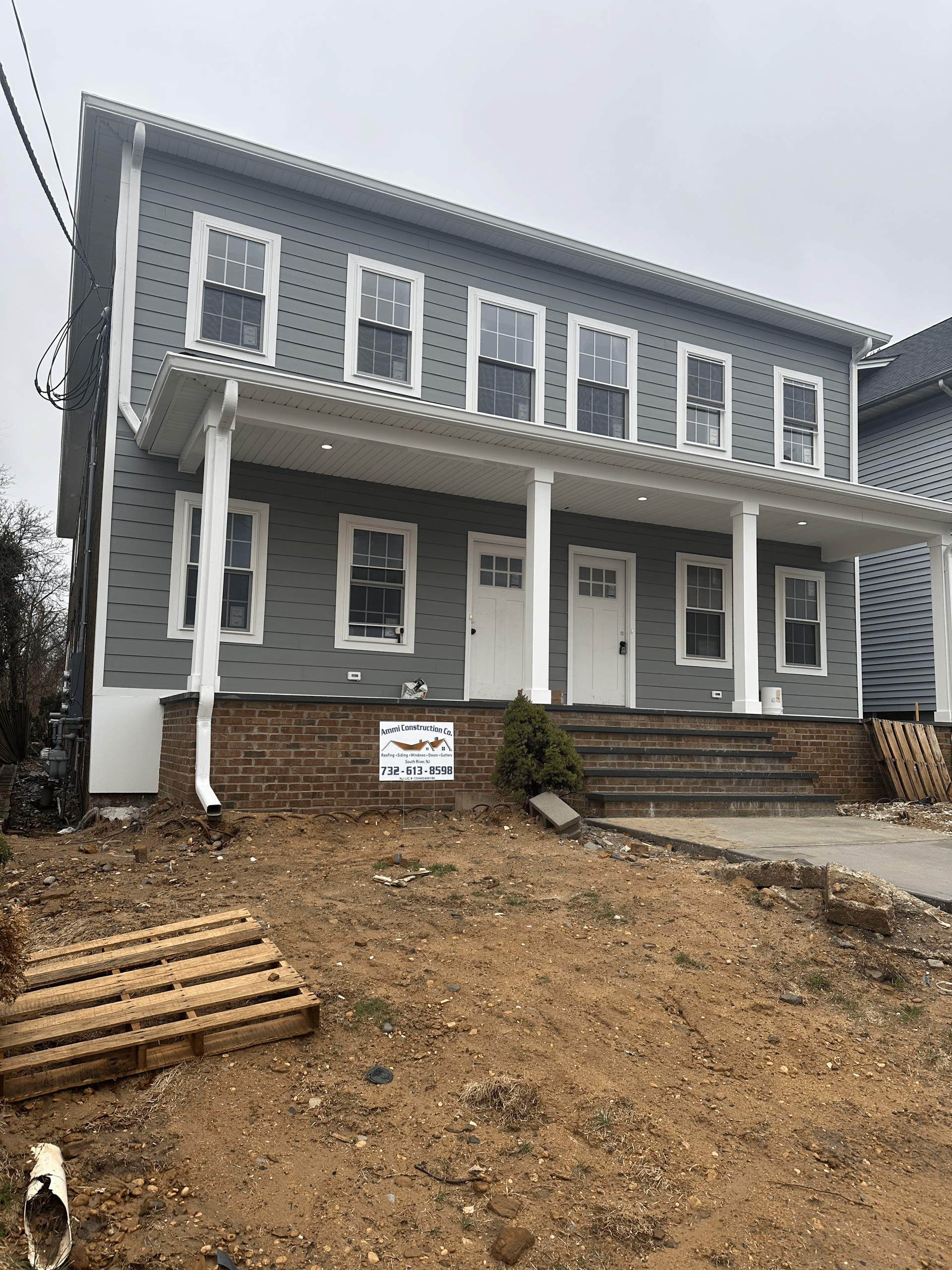 A two-story grey house under construction with a front porch, columns, and a dirt yard with a pallet on the ground.