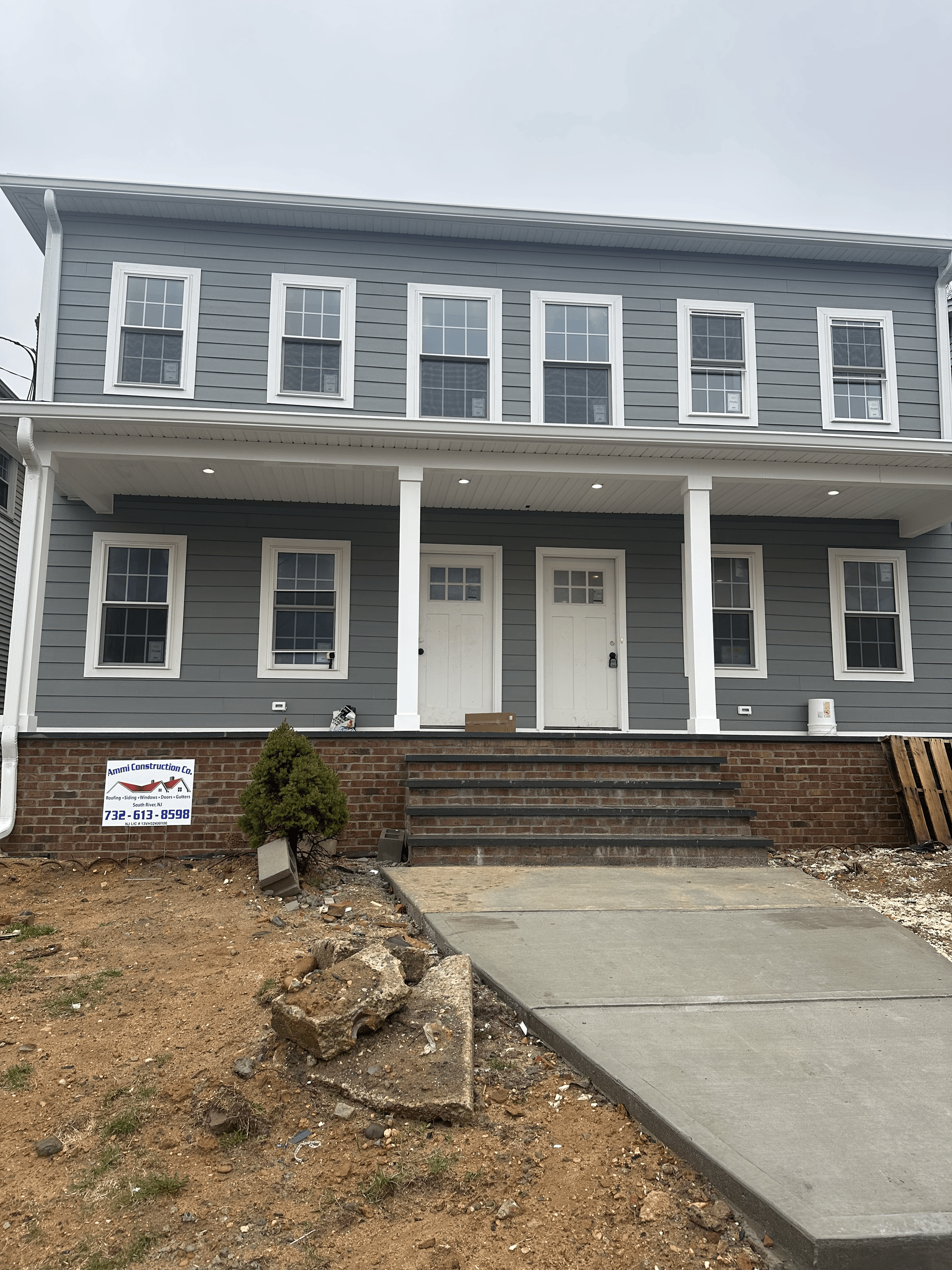 A two-story, gray house with white trim, a covered porch, and two front doors, surrounded by unfinished landscaping.