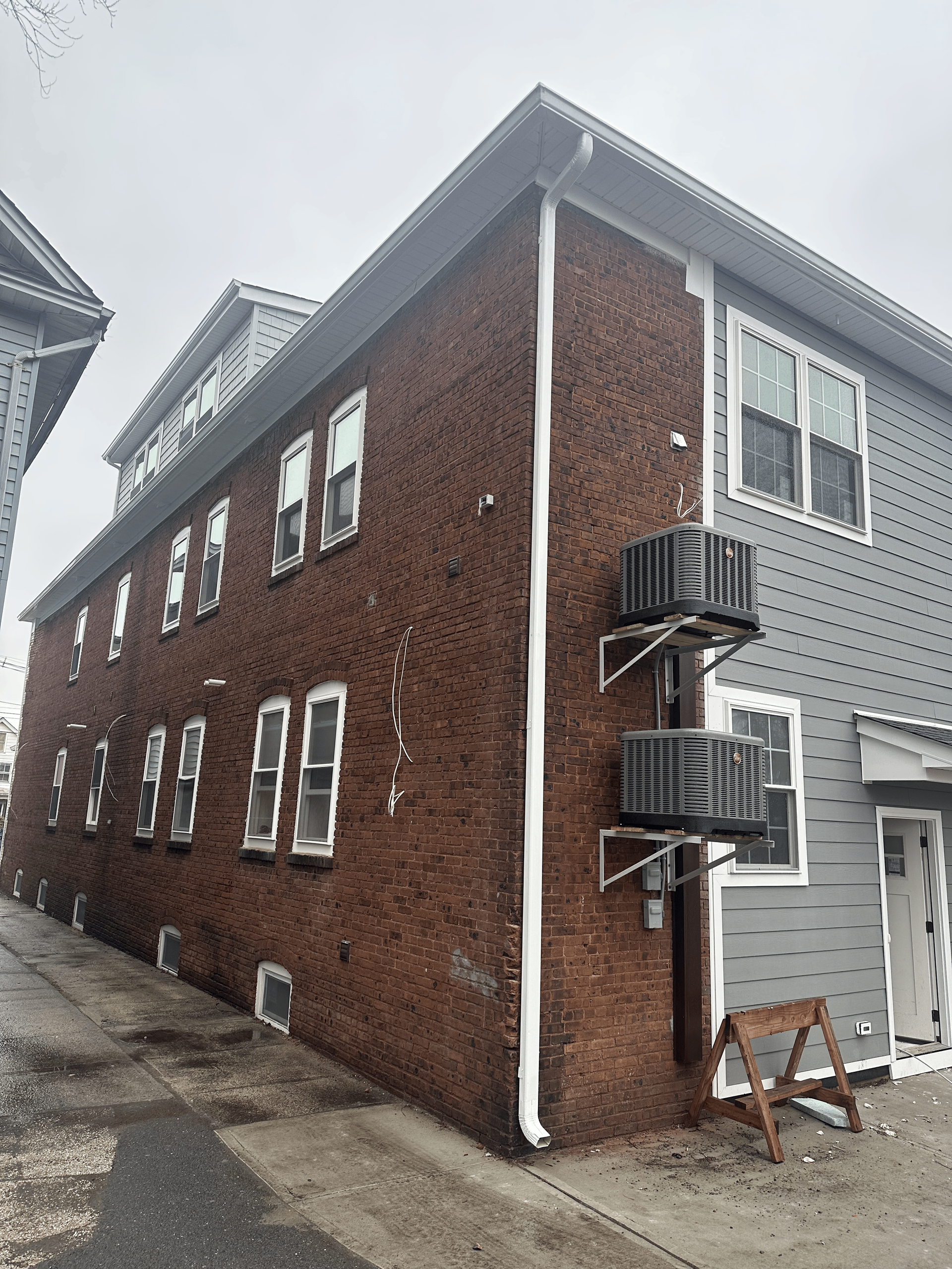 A building with a brick side wall featuring several windows, transitioning to grey siding on the front with two AC units.