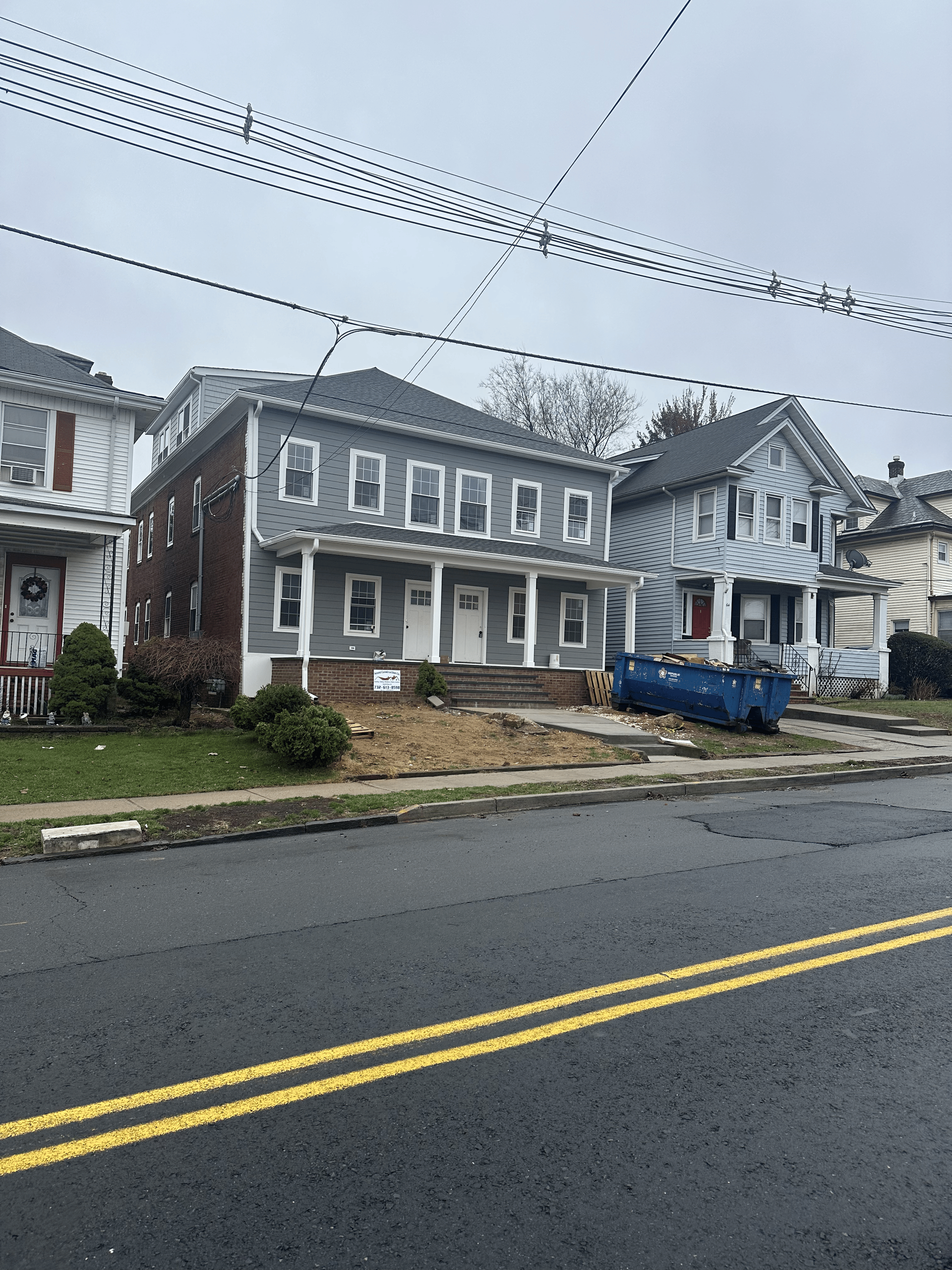 A grey two-story house with a blue dumpster in front, viewed from across a street with double yellow lines.