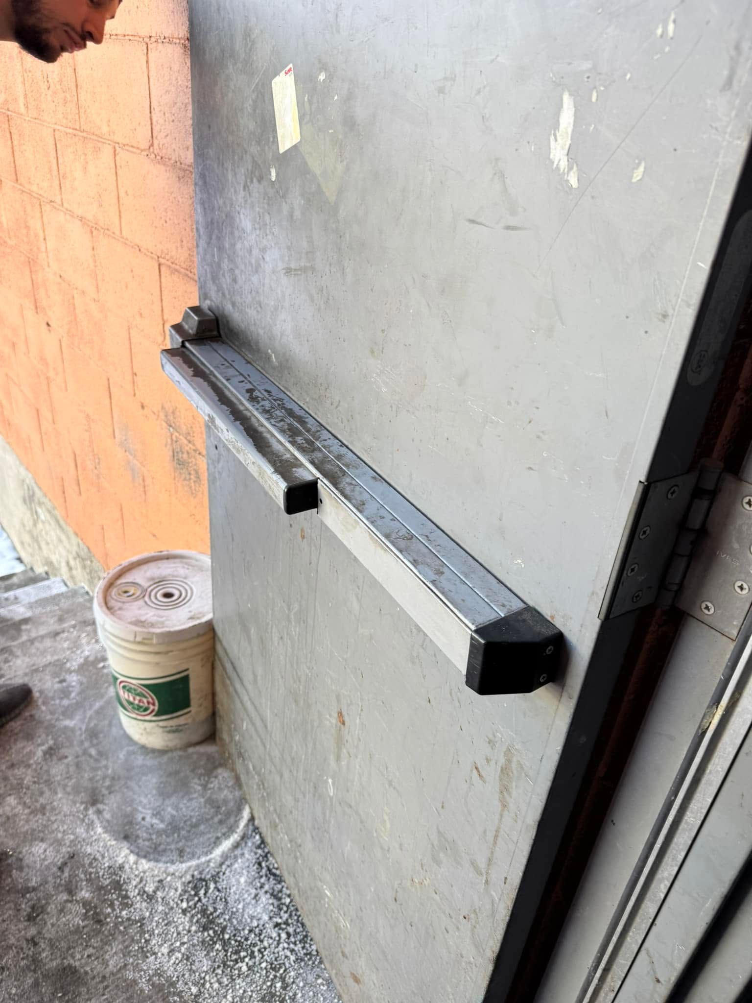 Wall-mounted metal barrier arm with a black knob on a gray door beside an orange wall and paint bucket.