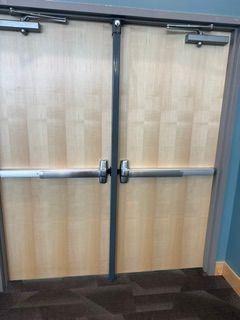 Wooden double doors with metal push bars in a hallway, viewed from inside.