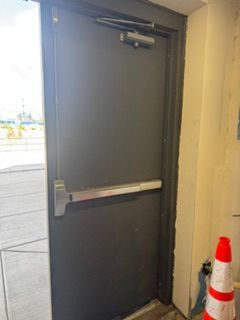 Closed gray metal door with push bar and overhead closer next to an orange traffic cone.
