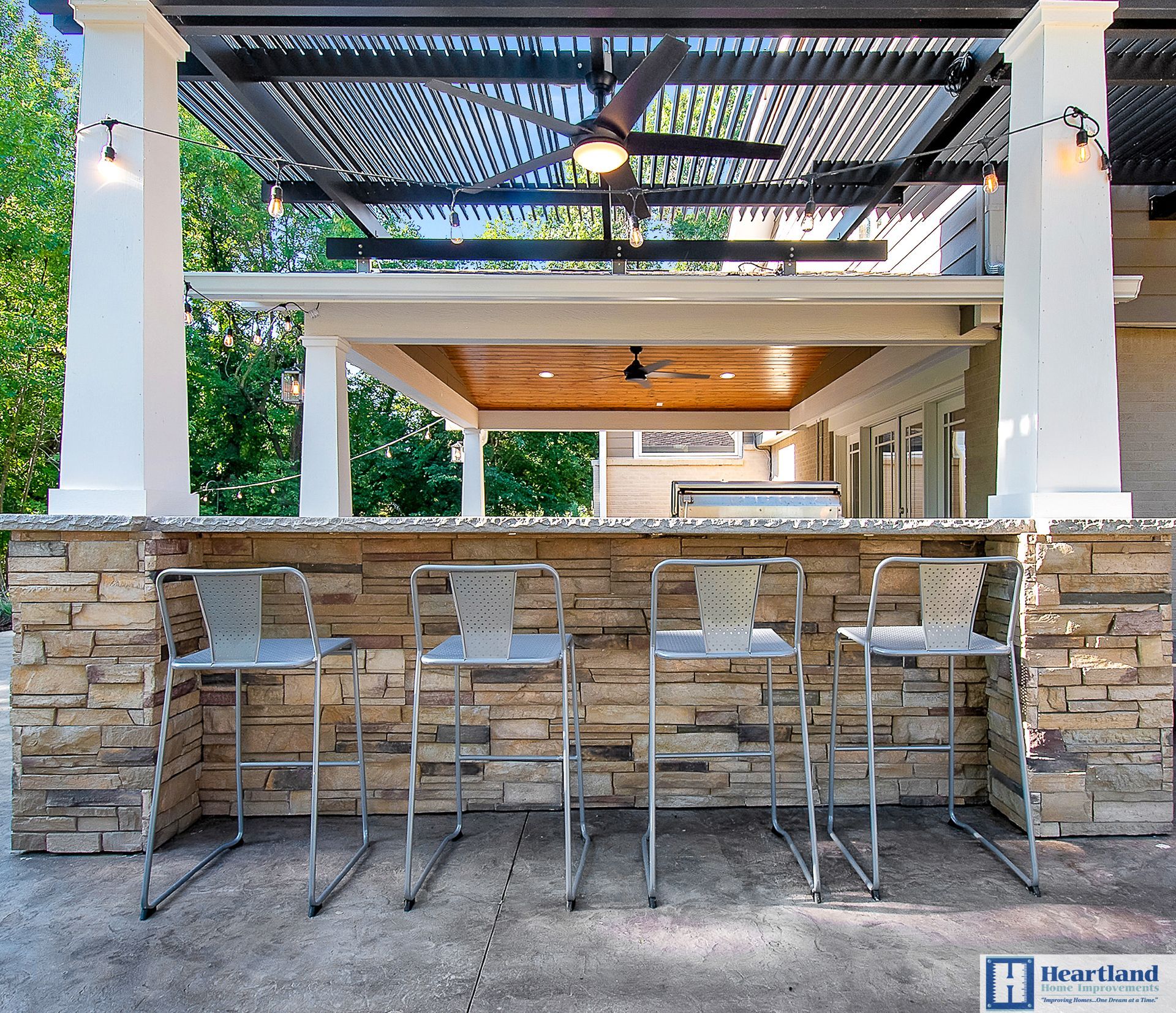 A stone bar with stools under a pergola and a ceiling fan