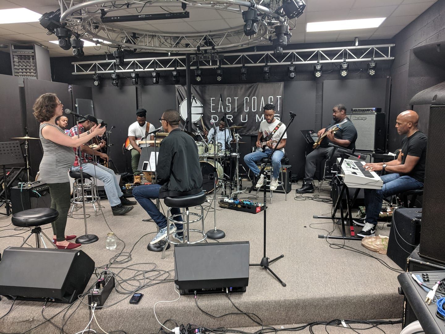 Band rehearsing on stage, with drummer, guitarists, keyboardist, and female director. Dark room with stage lighting.