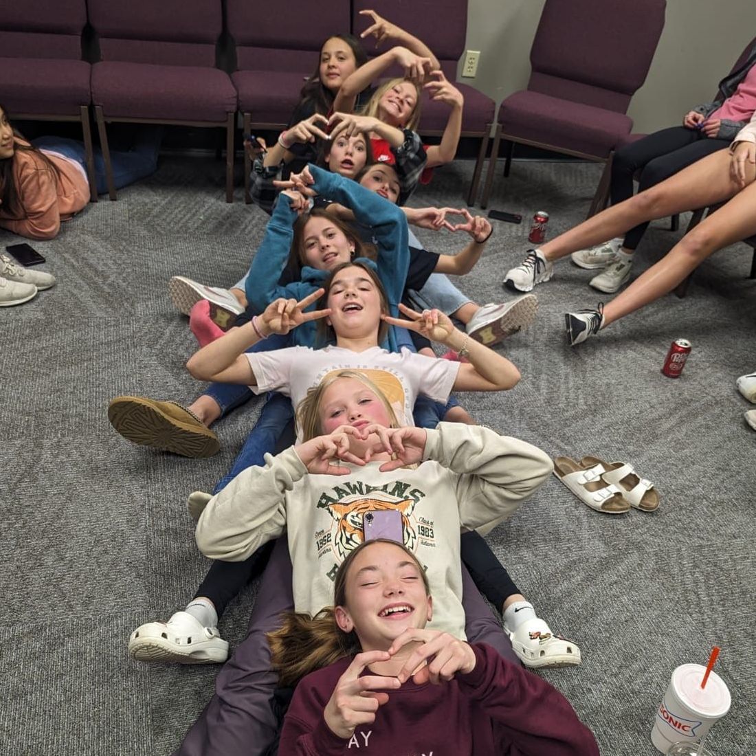 Group of young people forming a playful line, making hand gestures. They are on a carpeted floor near chairs.