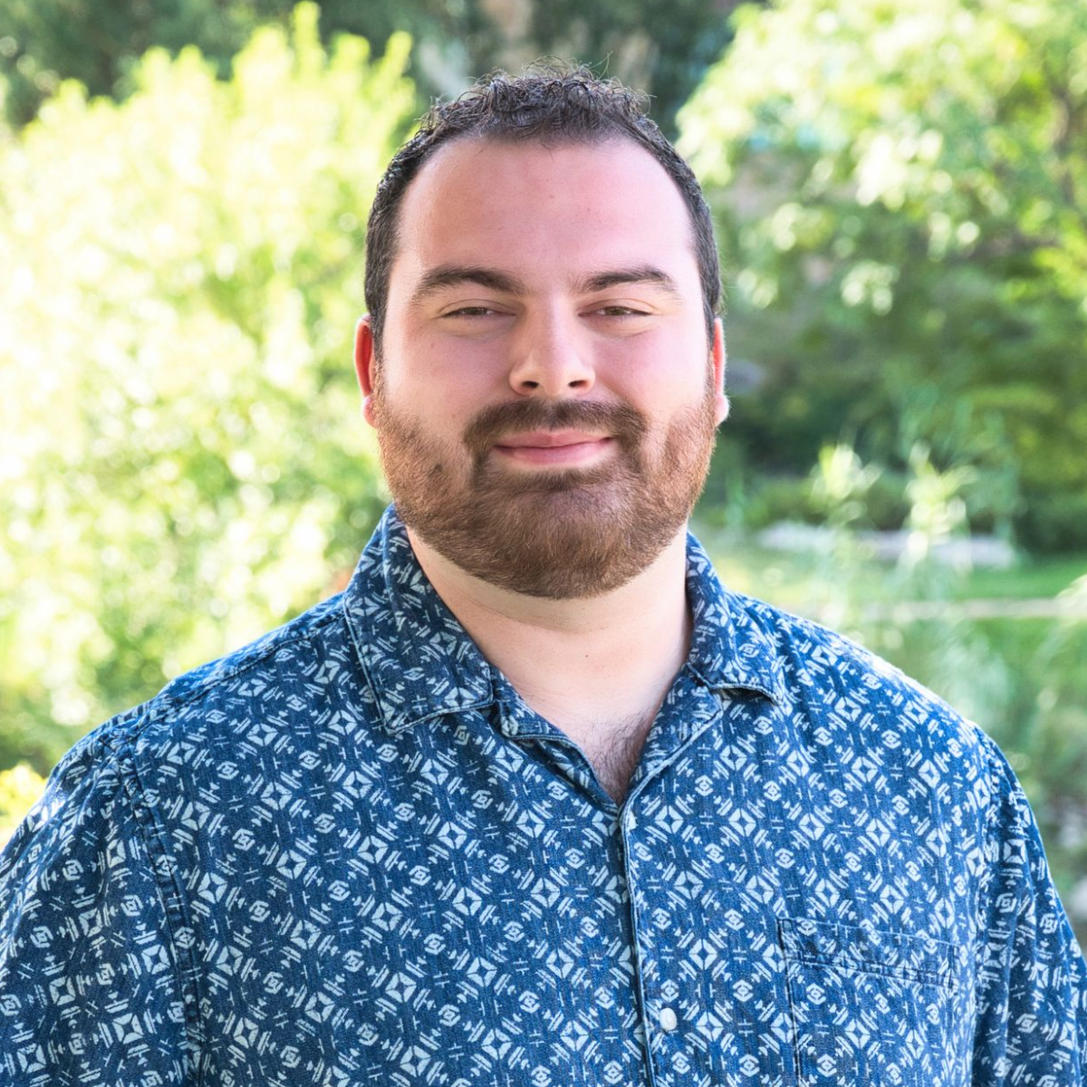 Man with long hair in a bun smiles while wearing a blue patterned shirt outdoors near a brick building.
