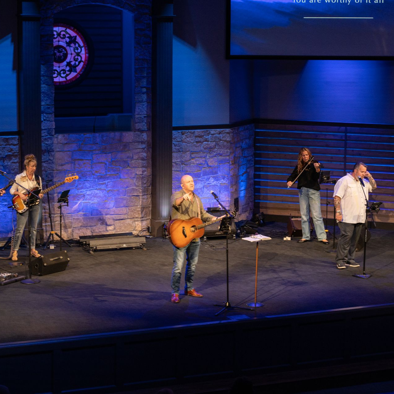 Band performing on stage in church. Musicians play guitar, bass, violin, and sing. Blue lighting.