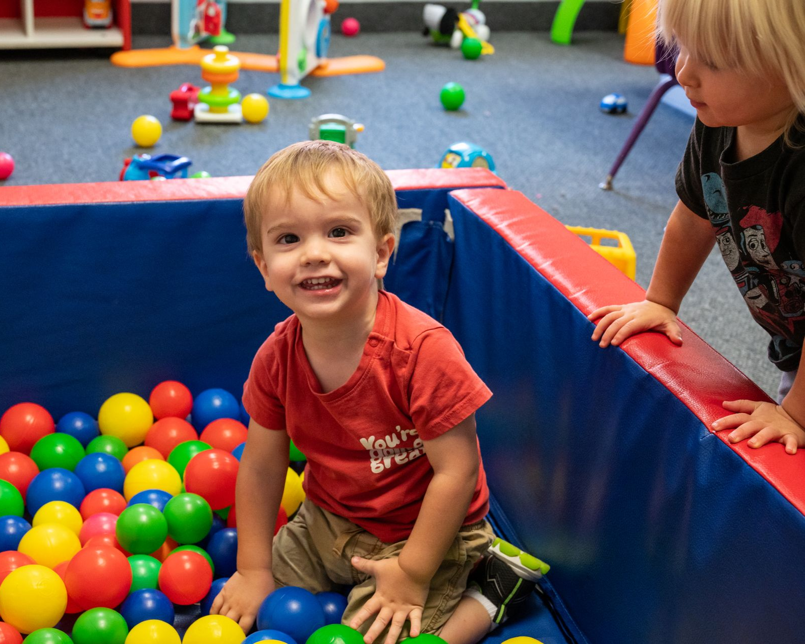 Boy smiling in a ball pit, another child nearby. Colorful balls, playroom setting.
