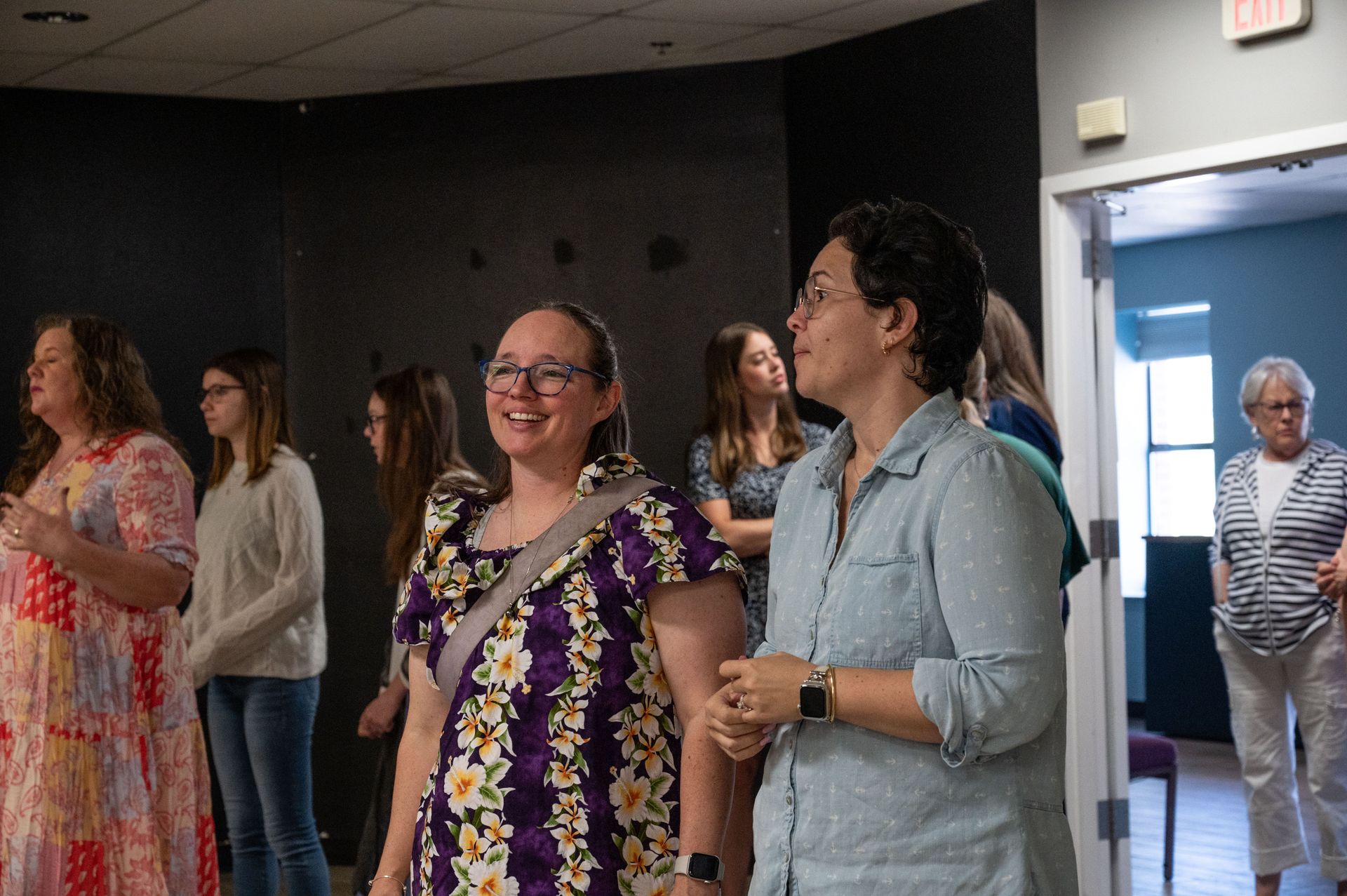 People standing and chatting in a hallway, with an open doorway and bright exit sign in the background.