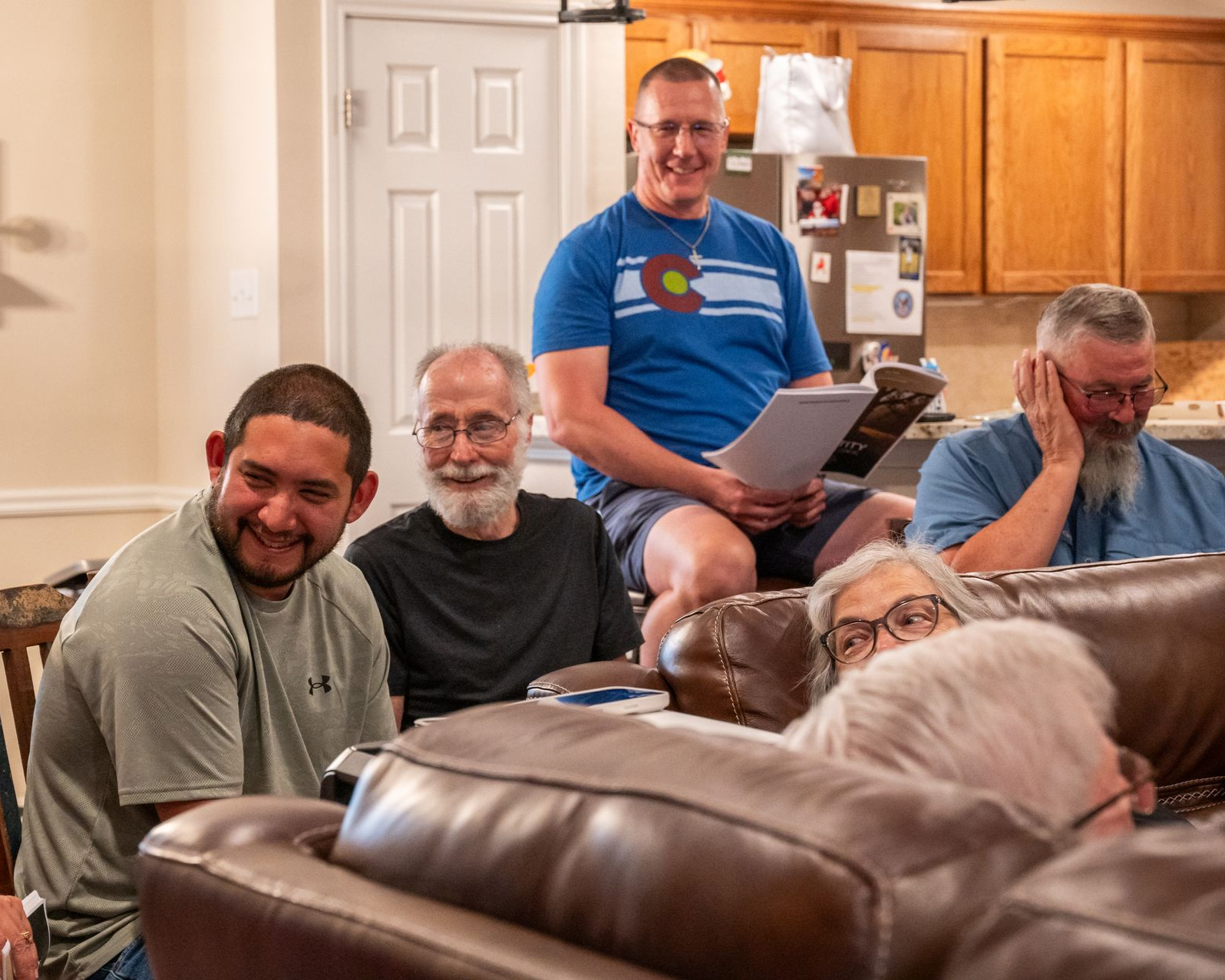 Group of people in a living room; one standing, reading; others sitting on couch, smiling and listening.