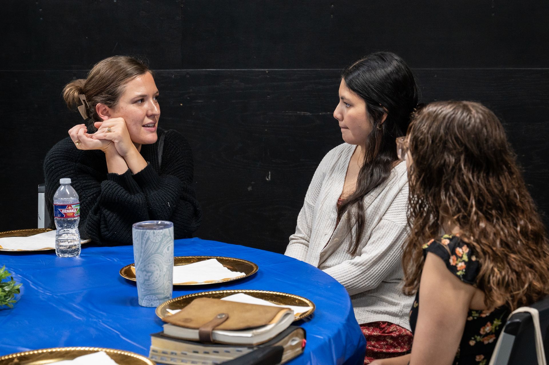 Three women talking around a table with a blue tablecloth at an indoor event