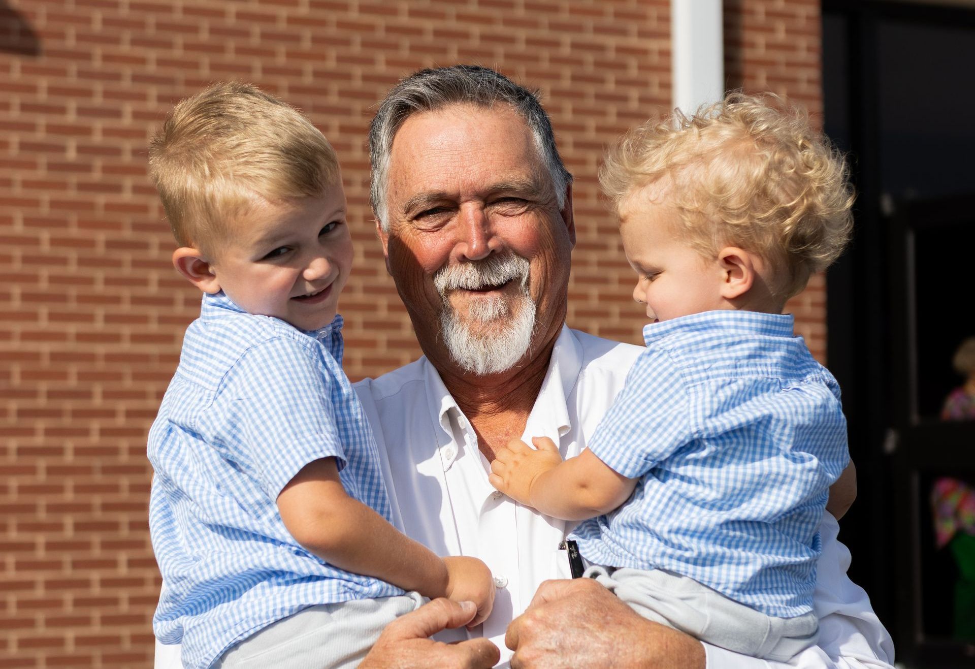 Man holding two young children, all smiling, outdoors in front of a brick building.