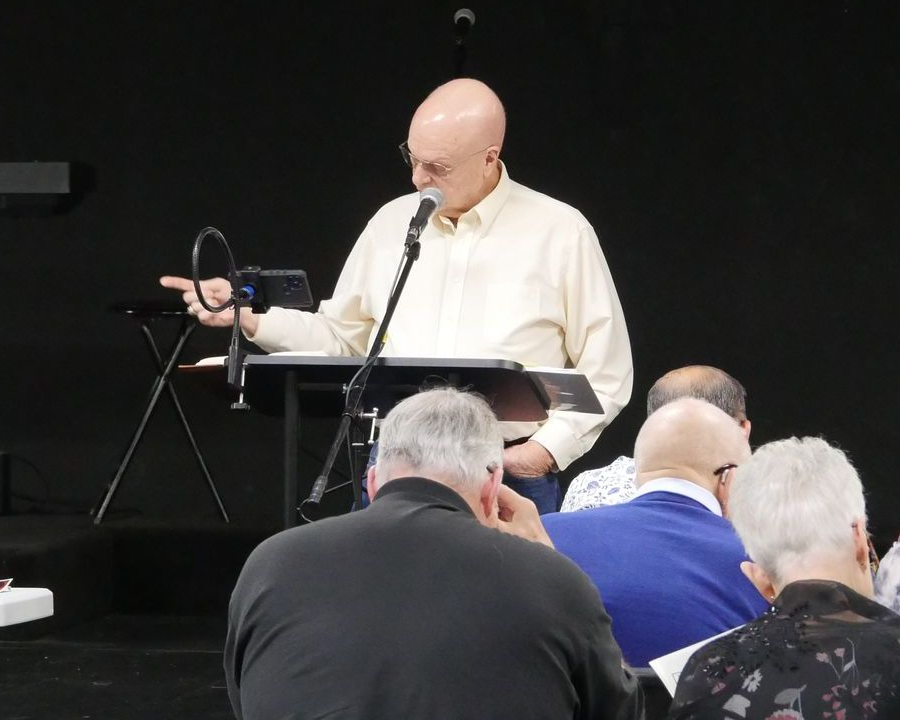 Man speaking into microphone, gesturing with ring. Audience sits in rows, listening.