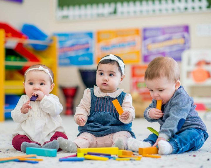 Three babies sitting on a rug playing with blocks in a colorful room.
