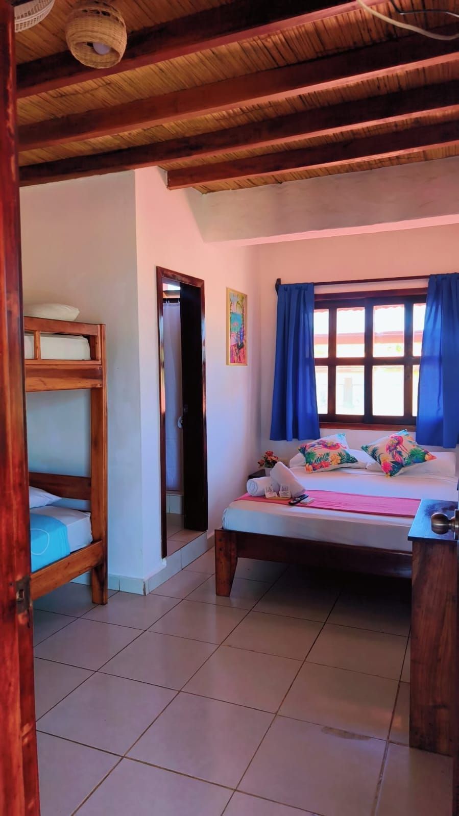 Cozy bedroom with a bed, blue curtains, tiled floor, and exposed wooden ceiling beams.