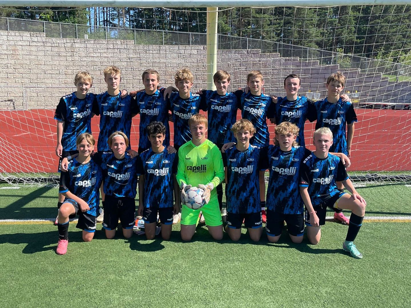 Soccer team in blue jerseys, kneeling and standing on a green field, goalkeeper in neon green.