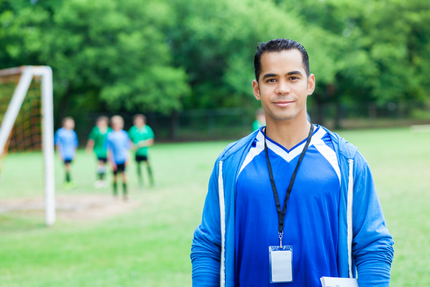 Soccer coach wearing blue stands on field, looking towards the camera; players in background.