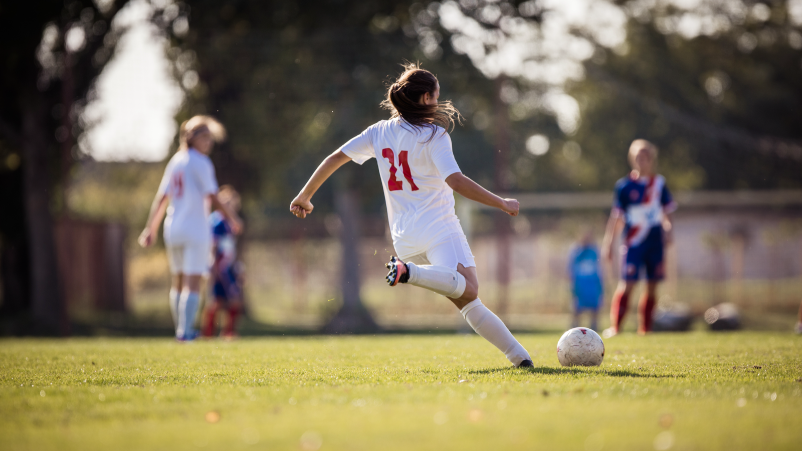 Soccer player in white uniform kicks a ball on a green field. Other players watch.