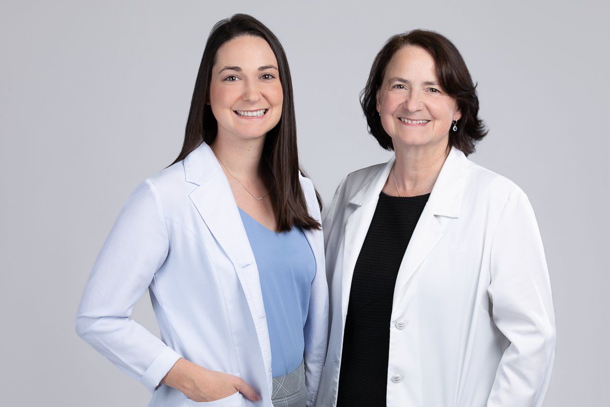 Two women in white lab coats smiling. One wears a blue top, the other a black top. Studio shot.