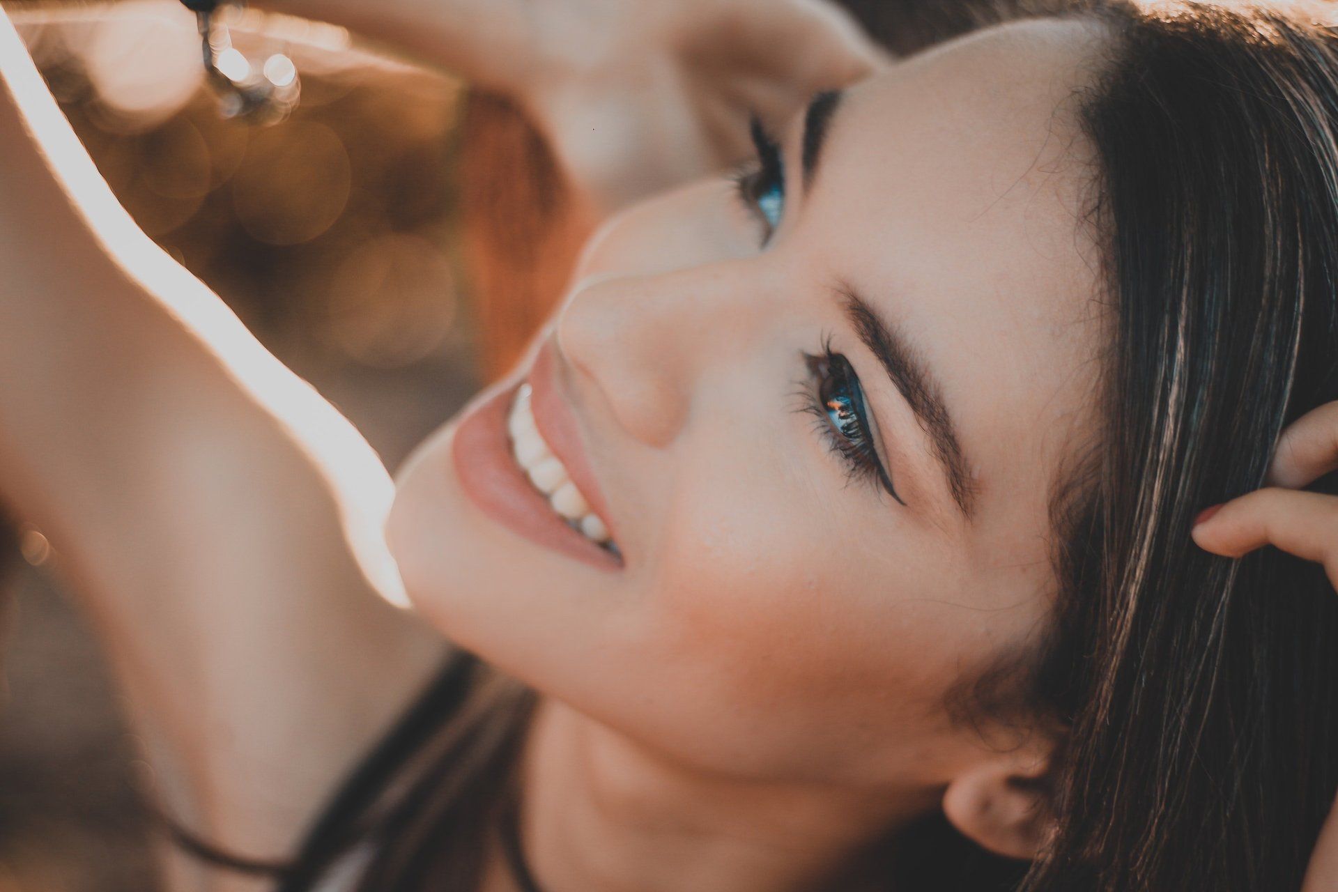 Une femme aux yeux brillants sourit, le regard tourné vers le ciel, les mains dans les cheveux, à l'extérieur, au soleil.