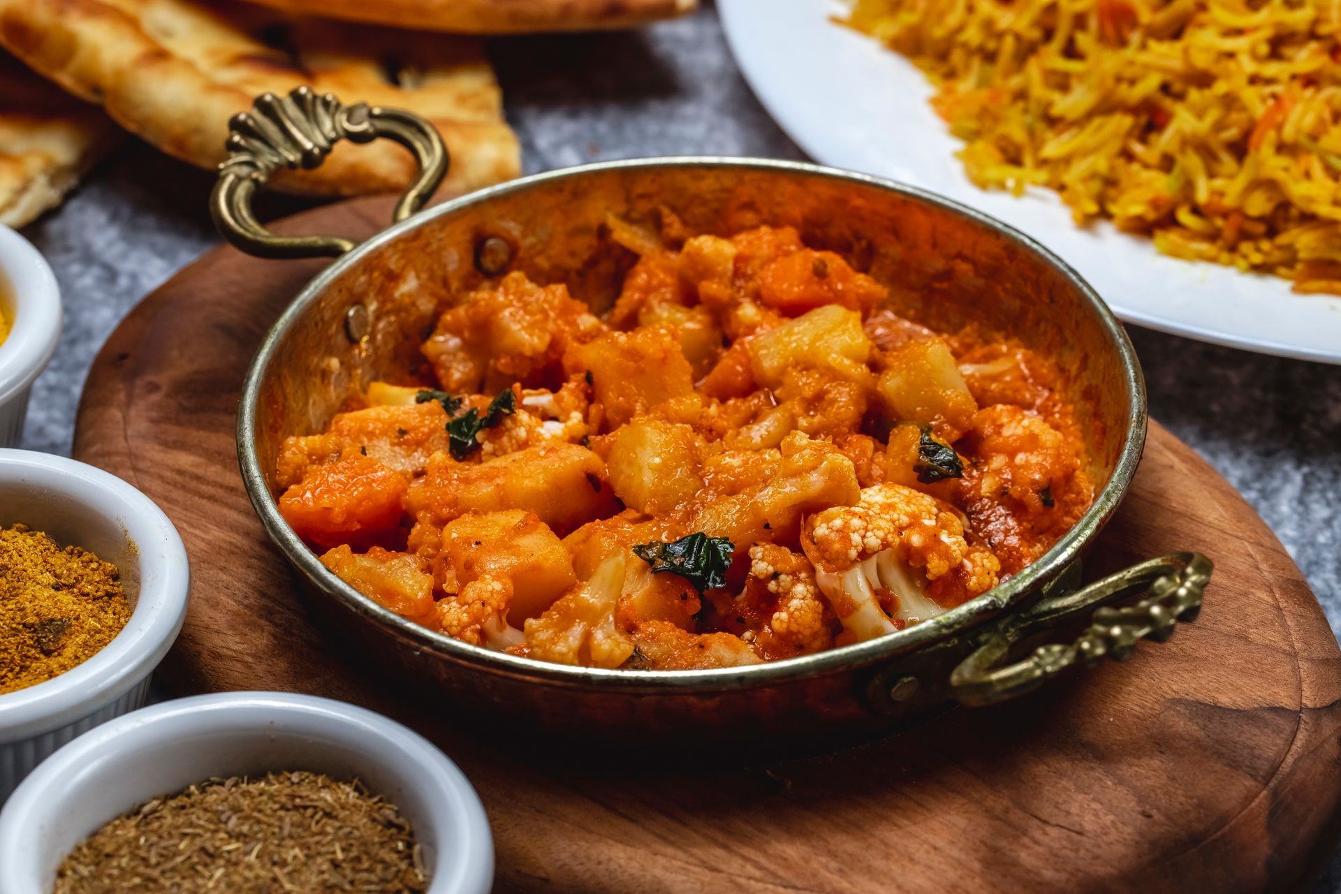 A copper bowl of aloo gobi on a wooden board, with side dishes of rice, naan, and spices on a grey surface.