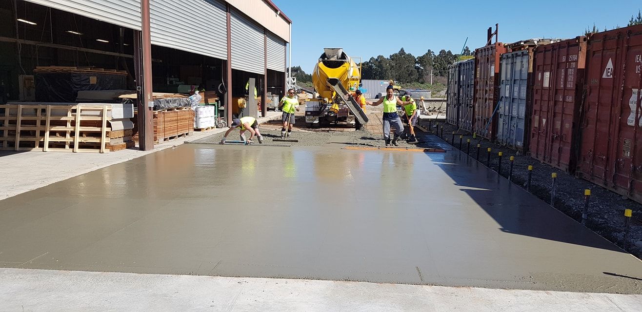 Workers pouring concrete with a truck. They are smoothing it out near a building and containers.