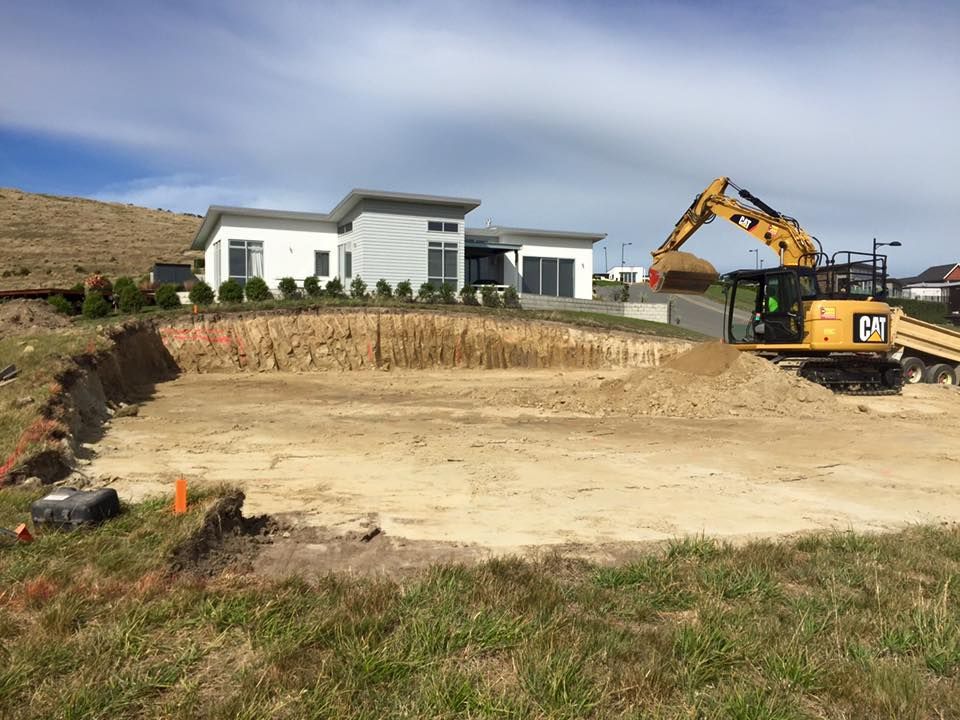 Excavator digging a foundation next to a modern white house on a sunny day.