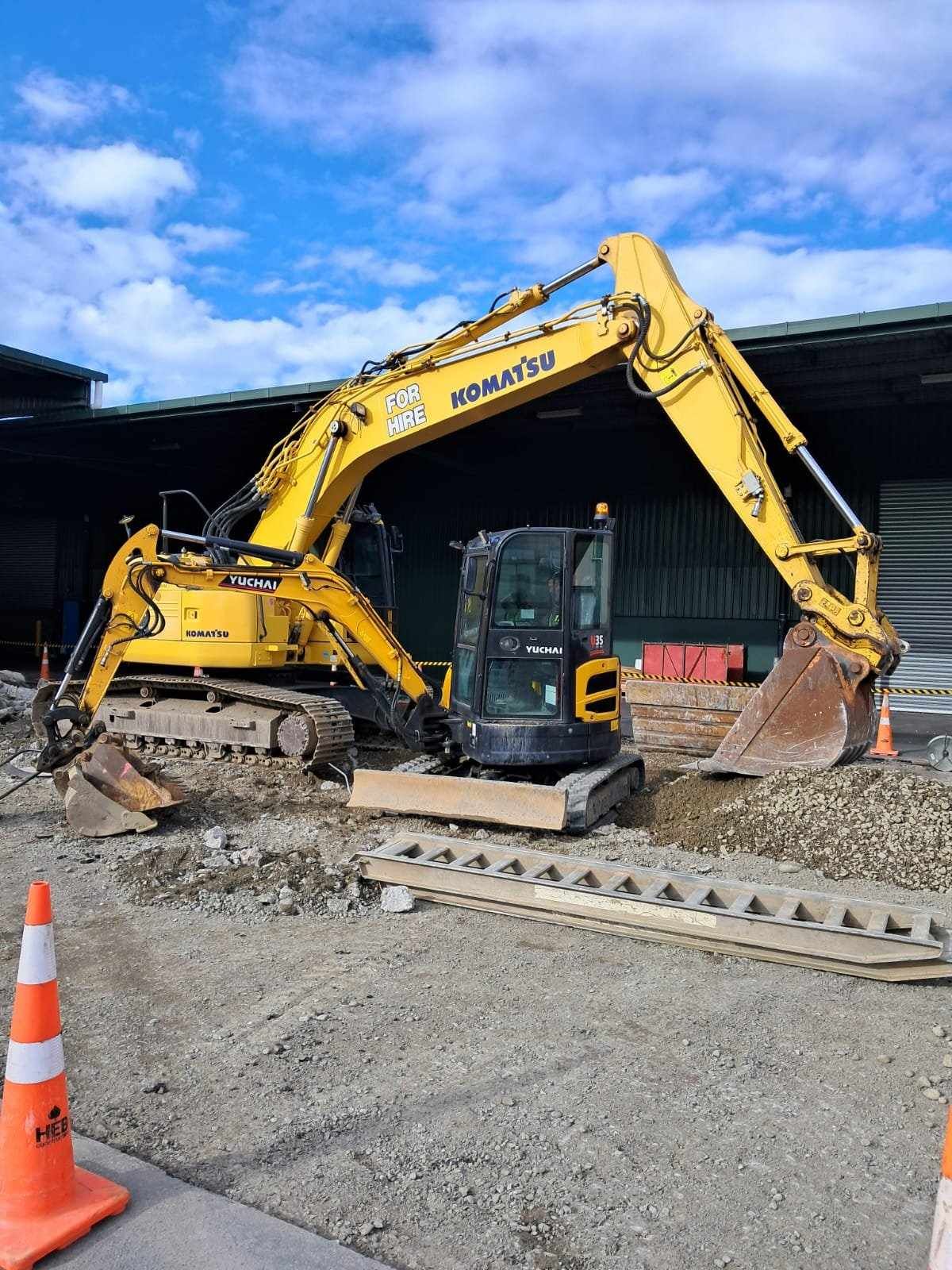 Yellow Komatsu excavator on gravel, with arm raised, beside a smaller excavator. Blue sky background.