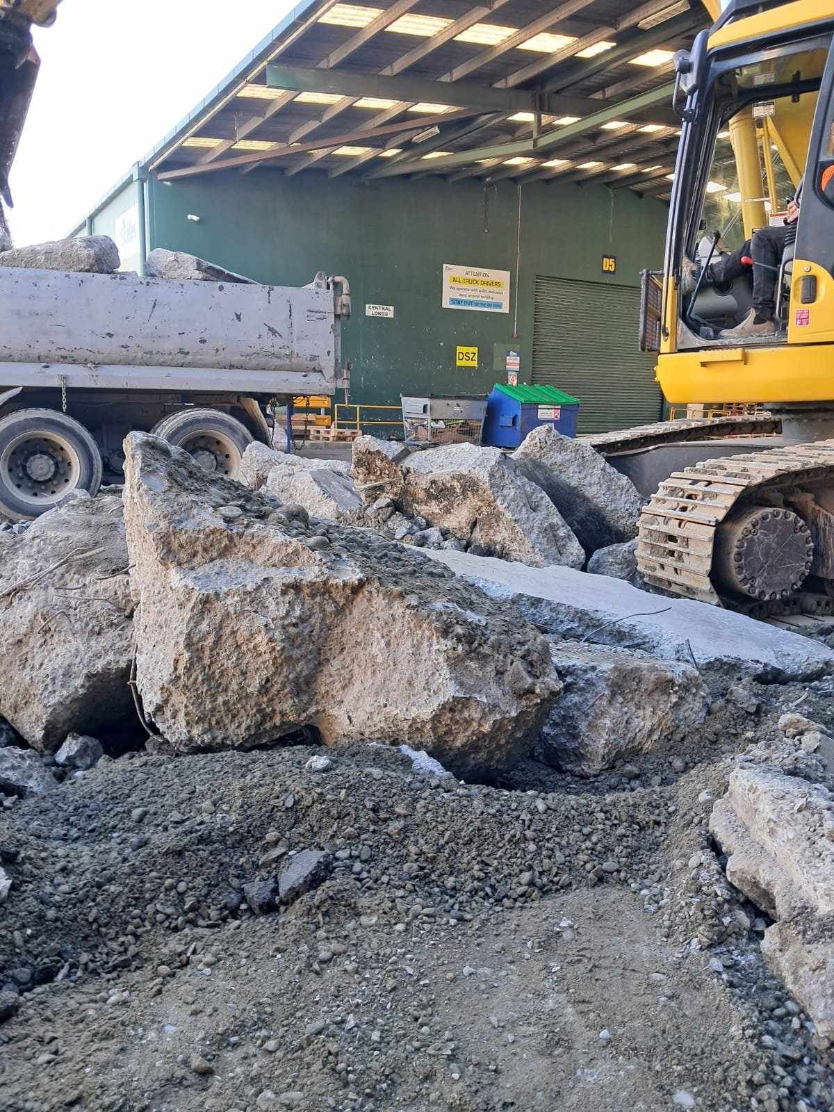 Construction site with excavator loading concrete debris into a dump truck, worker nearby.