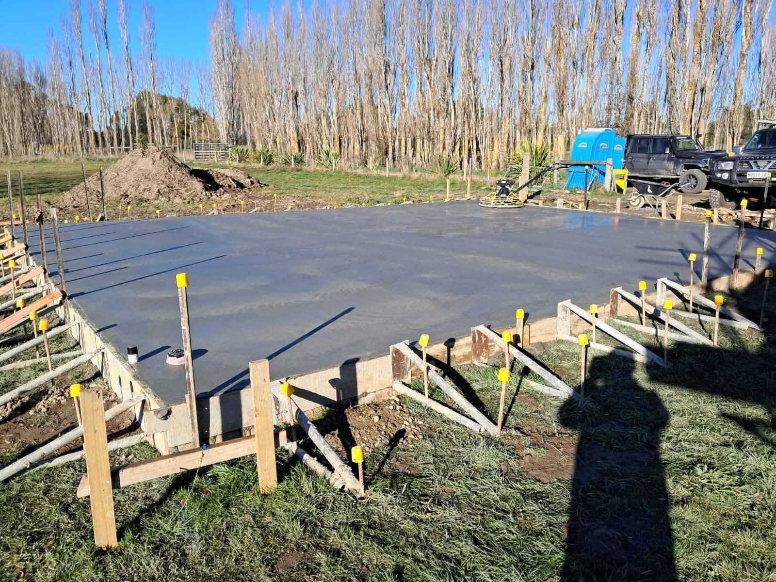 Freshly poured concrete foundation within wooden formwork, outdoors in a field, with trees in the background.