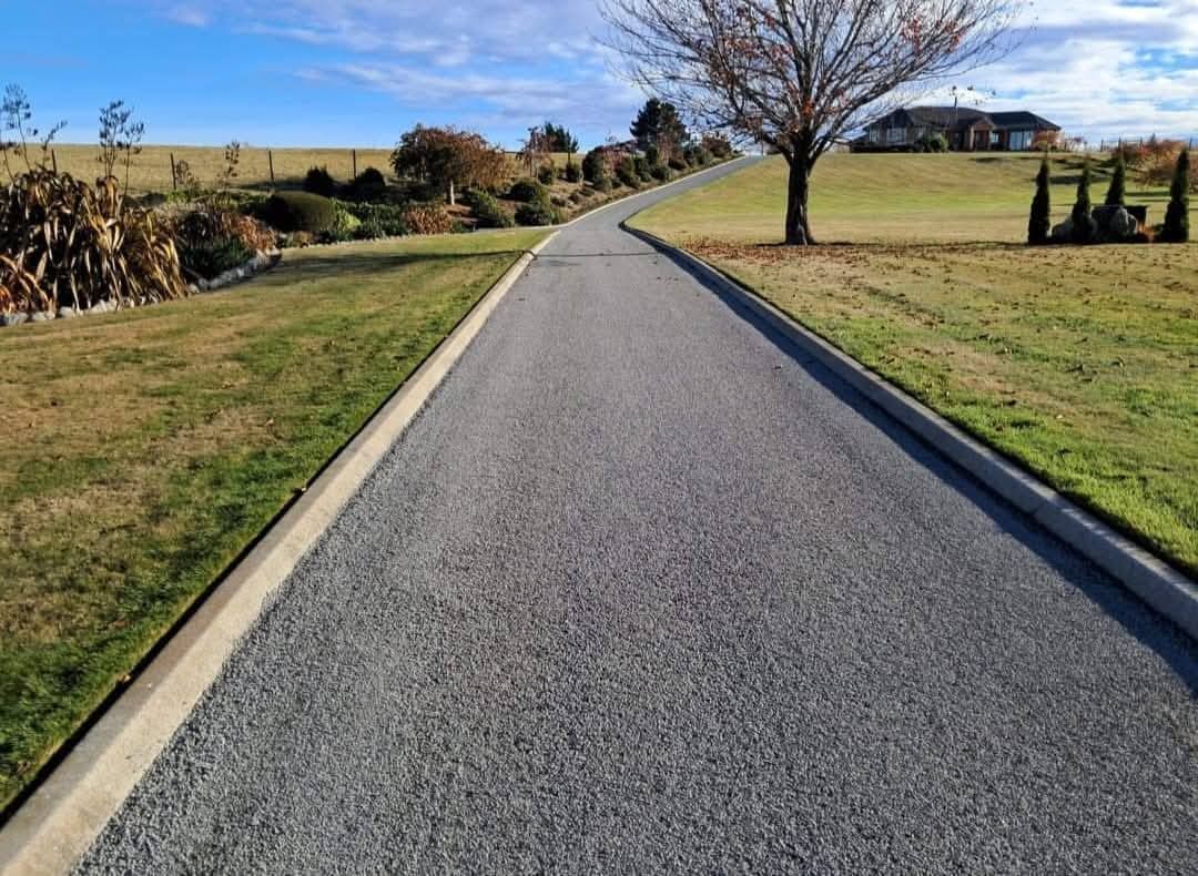 Asphalt driveway uphill, bordered by grass and concrete, leading to a house in the distance.