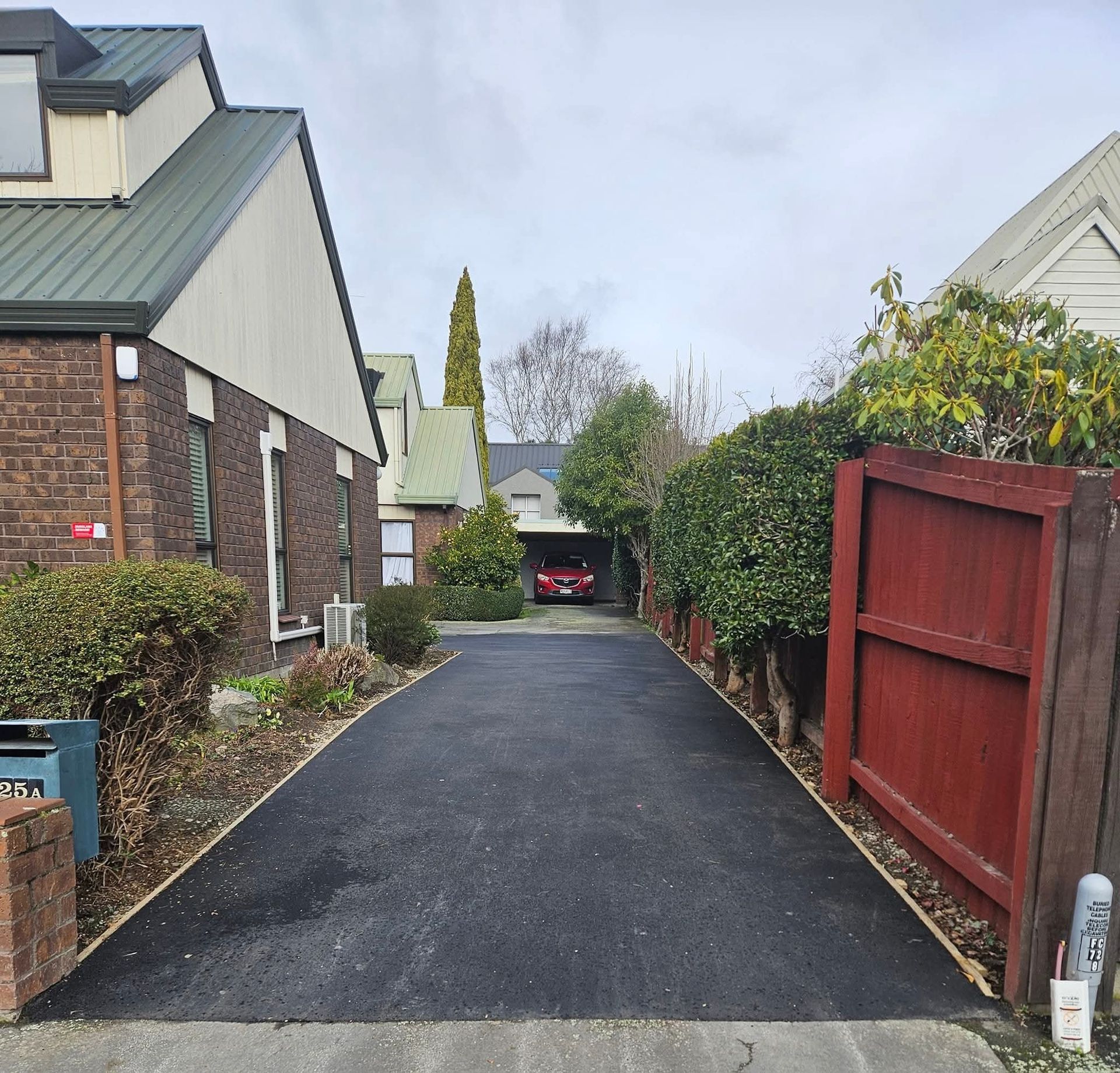 Black asphalt driveway leading to a garage with a red car. Houses line the sides with greenery.