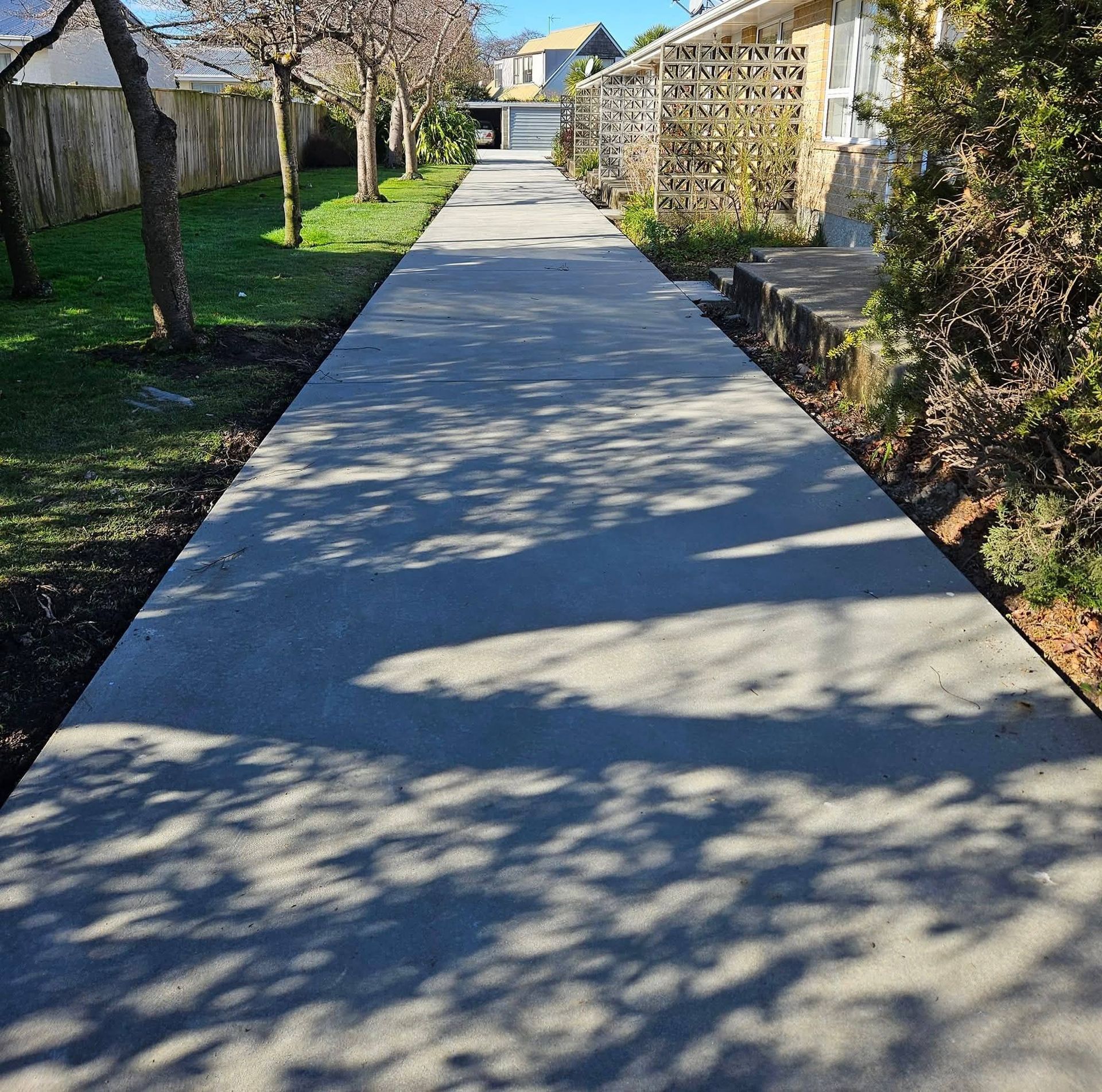 Concrete walkway with tree shadows, bordered by grass, trees, and houses.