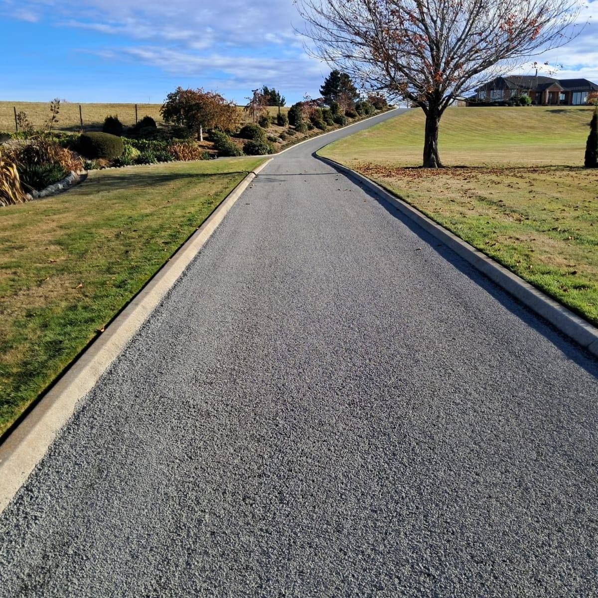 Gravel driveway curving uphill, bordered by grass and a concrete curb. A bare tree and a house are visible.