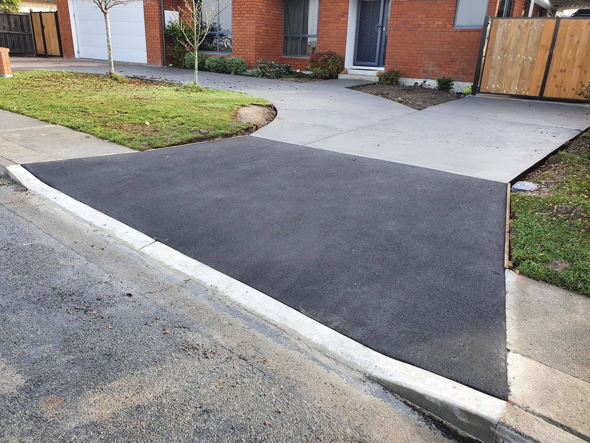 Asphalt driveway apron connects to a concrete driveway and sidewalk. Green grass borders. Brick house in background.