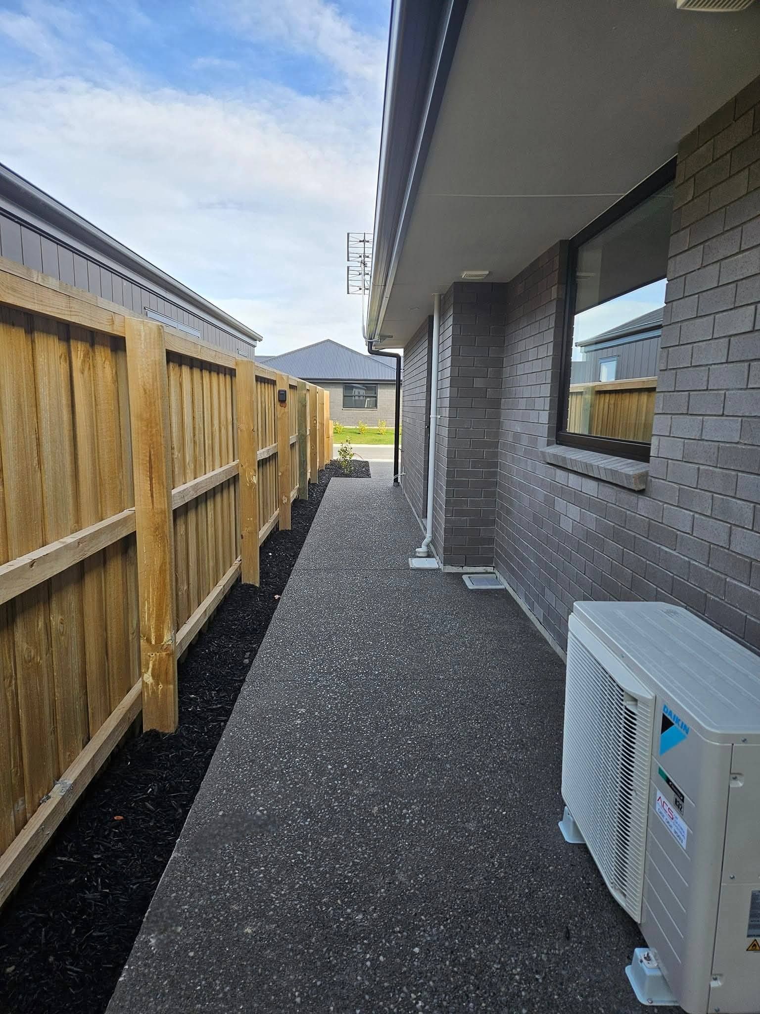 Narrow walkway between a wooden fence and a brick wall with an air conditioner unit.