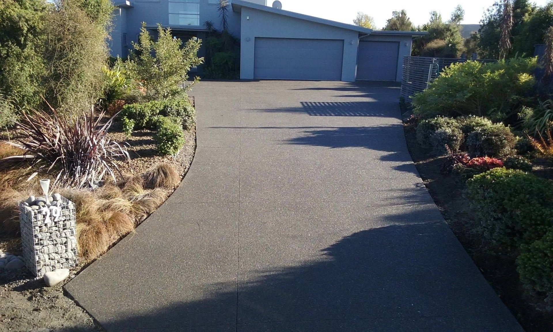Dark driveway leads to a gray house with a garage, flanked by lush landscaping and a rock feature.